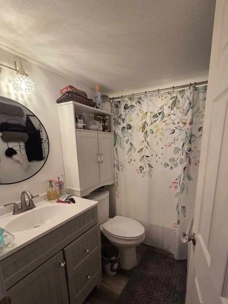 A small bathroom with a light-colored vanity, a round mirror, a toilet with a floral shower curtain, and a wall cabinet. There are various toiletries on the sink and a small dark trash bin below the cabinet.