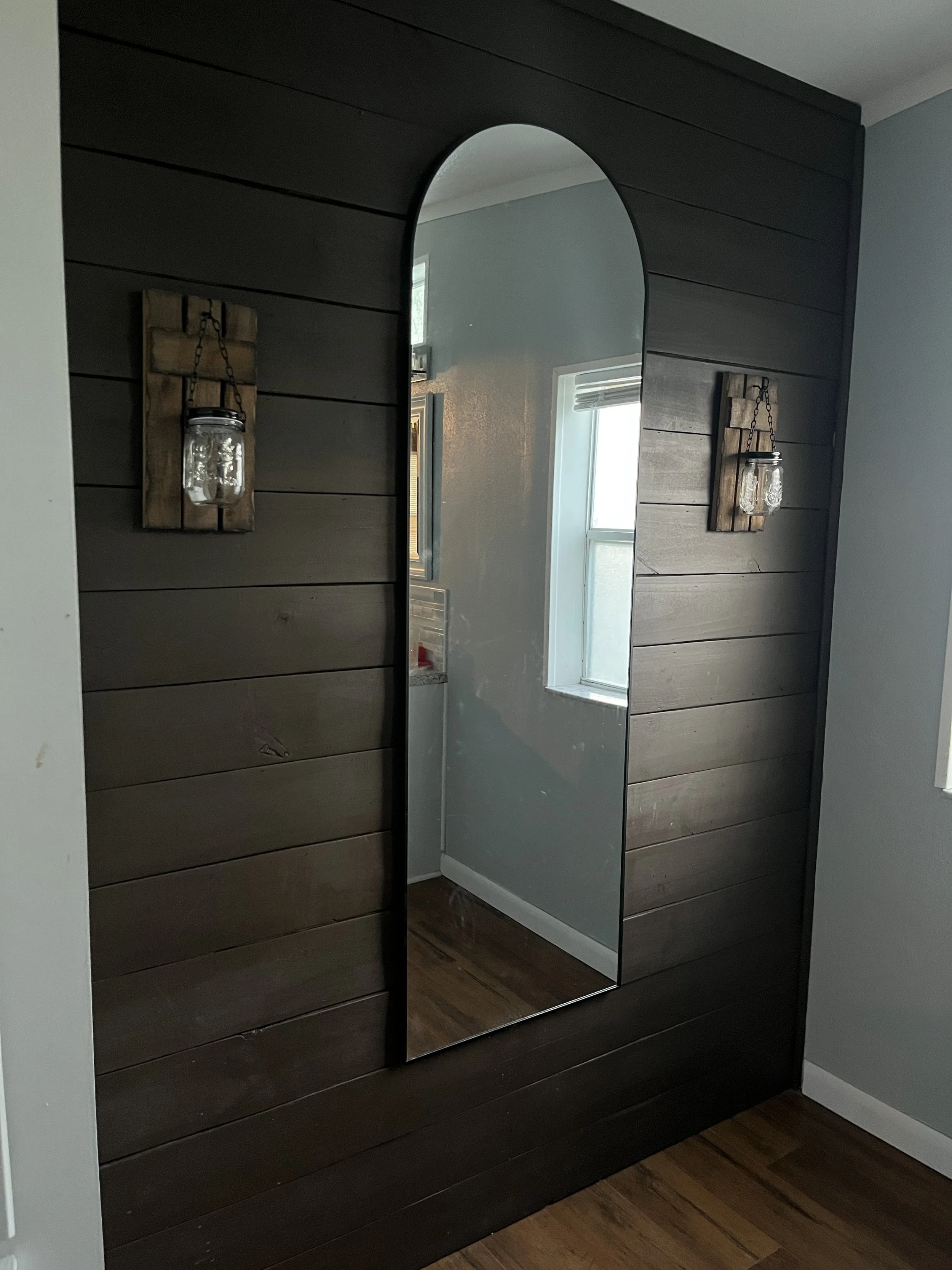 A tall mirror with an arched top mounted on a dark wooden wall panel, with a wall sconce featuring a Mason jar style glass shade with decorative design on each side.