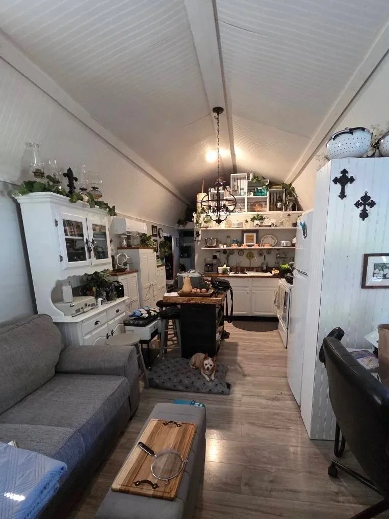 A cozy kitchen with open shelves, white cabinets, and a rustic dining table. There is a gray couch, a small dog sitting on a mat, and a chandelier hanging from the ceiling. The room features wood flooring and decorated shelves with plants and kitchenware.