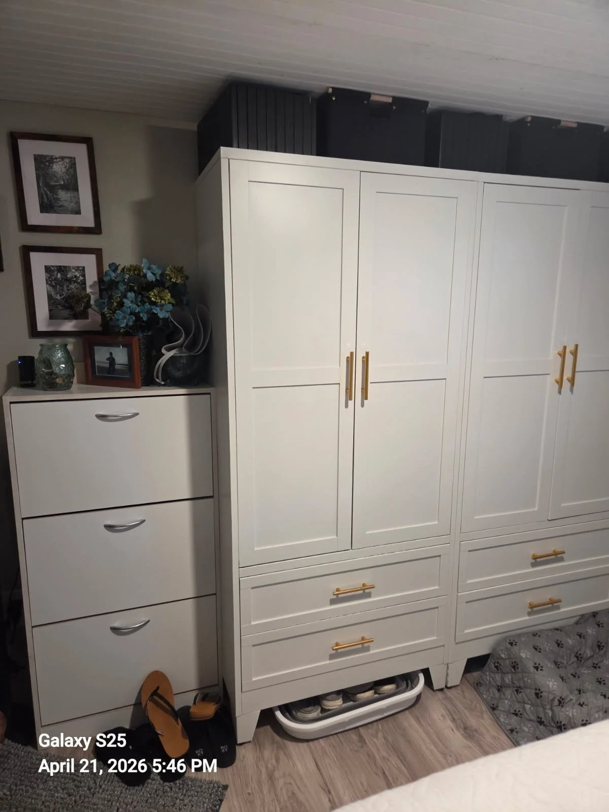 A tall white wardrobe with gold handles next to a smaller white dresser with three drawers, decorated with picture frames, a vase with flowers, and a small speaker. Shoes are on a tray beneath the wardrobe, and a patterned blanket is partially visible on the right side of the image.