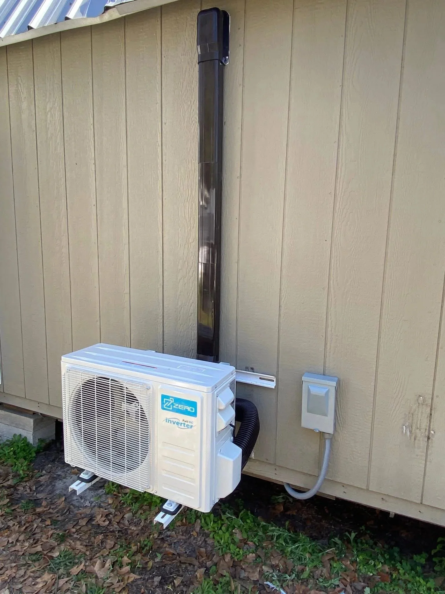 An outdoor air conditioning unit installed on the ground, connected to a house wall with a metal vent pipe. The wall is beige with vertical siding, and there is a small white electrical box nearby. The surroundings include some green plants and fallen leaves.