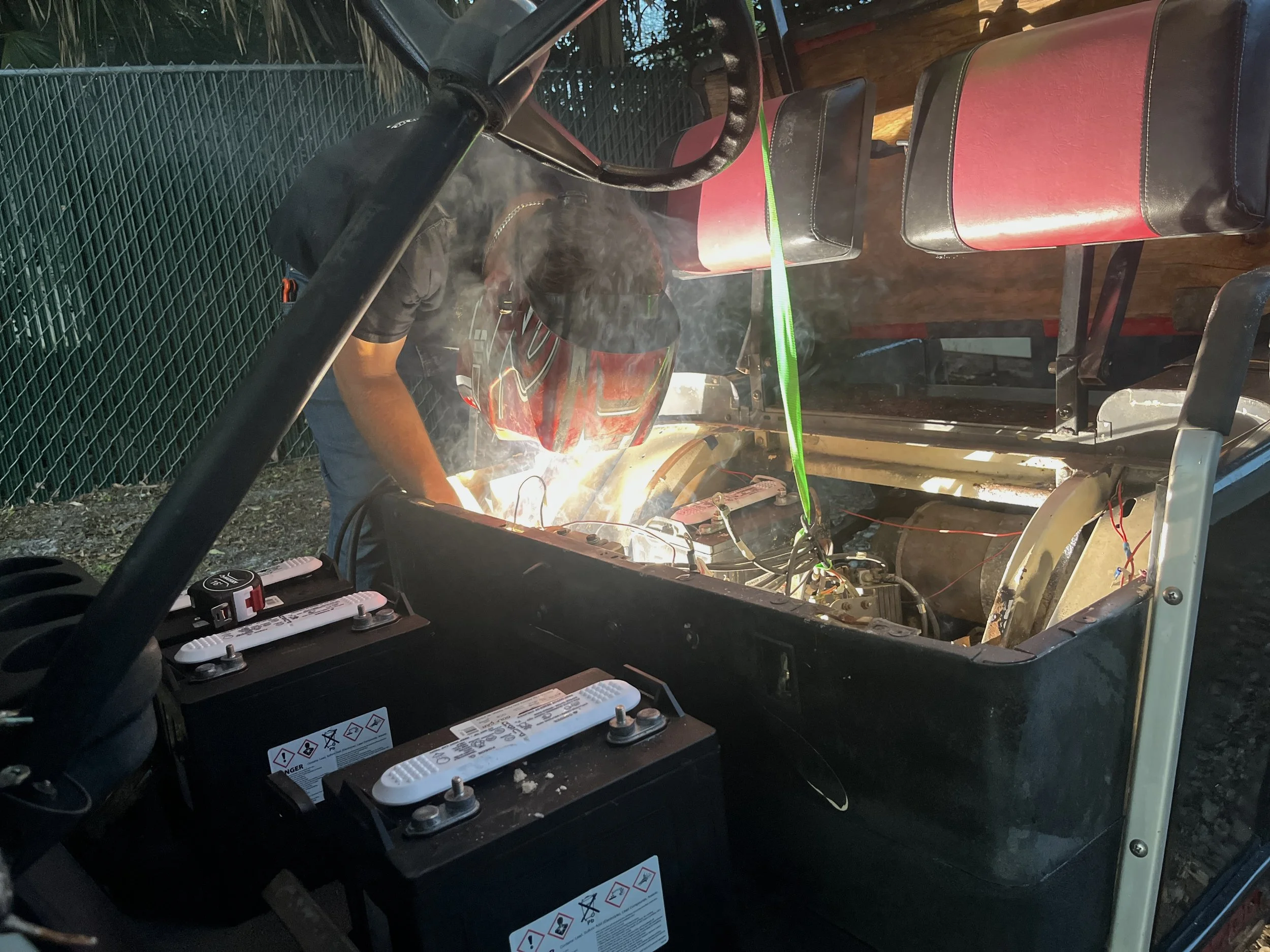A person working on a red vehicle engine outdoors, with green trees and a wire fence in the background. The person is wearing black and is welding or repairing the engine, which has smoke and sparks around it. The image shows the interior of the vehicle with red and black seats.