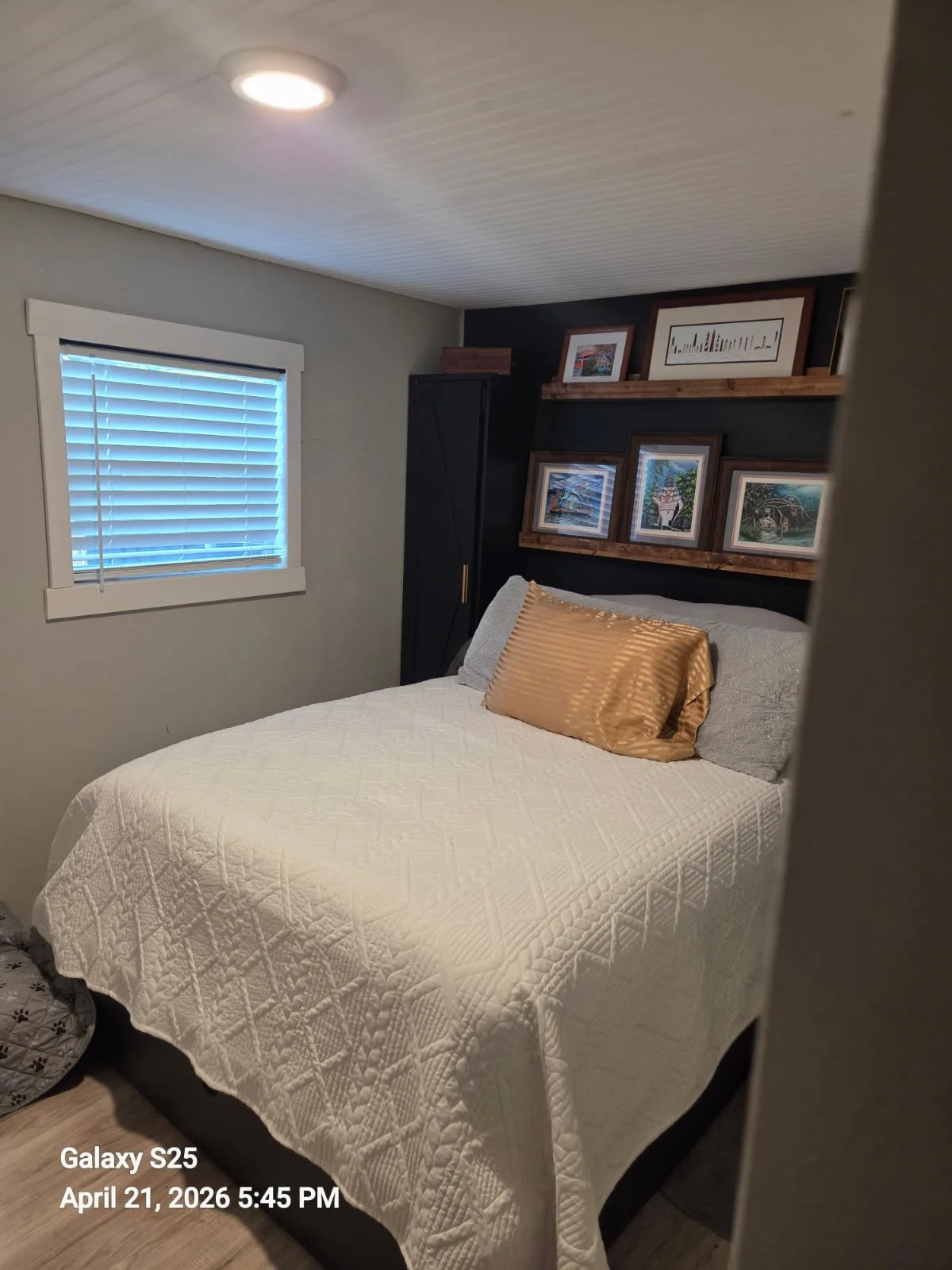 A cozy bedroom with a white quilted bedspread, a gold satin pillow, granite gray pillows, a window with closed blinds on the left, and framed artwork on a dark accent wall above the bed. The room has wooden floors and a ceiling with white beadboard paneling.
