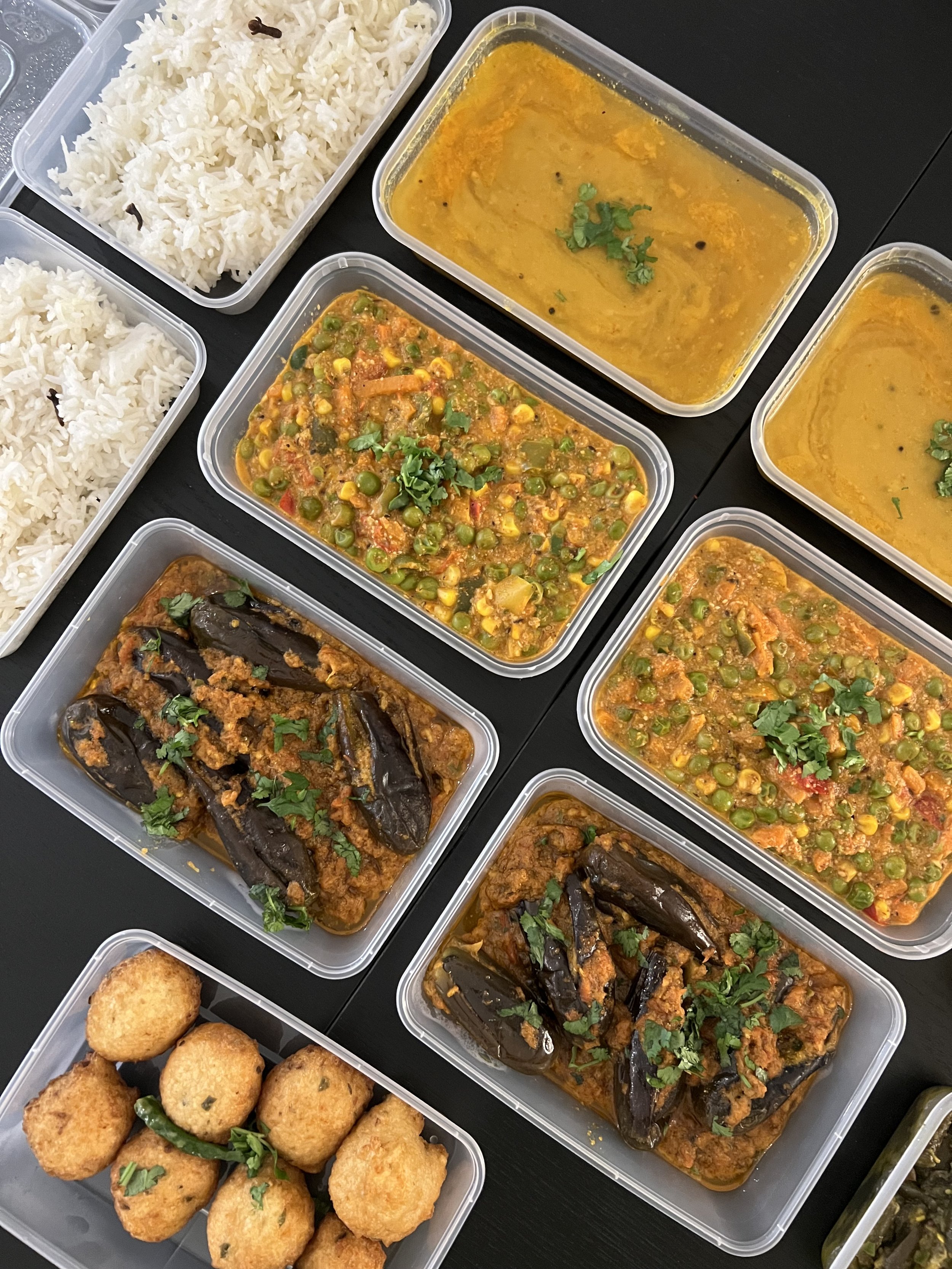 Meal with white rice, spicy vegetable curry with peas, corn, and tomatoes, and eggplant in a tomato-based sauce, all in plastic containers, with some fried snacks on the side.