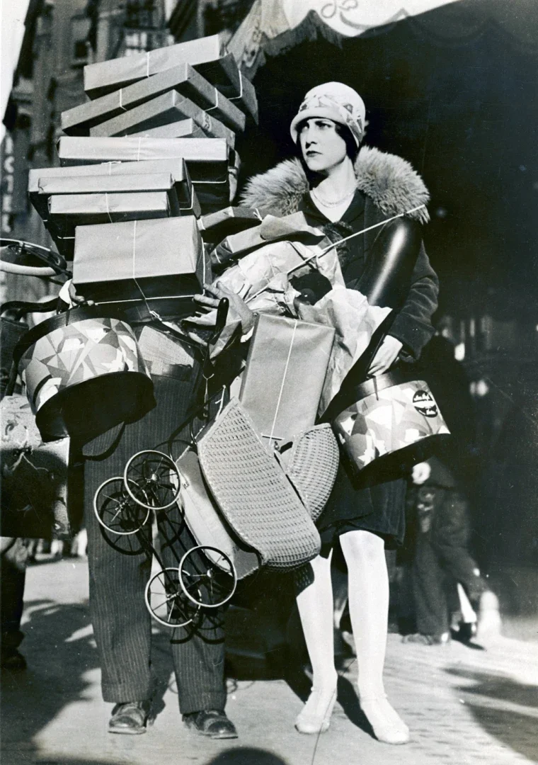 A woman in vintage clothing with a hat and fur collar shopping or commuting, carrying numerous packed boxes, bags, and a helmet, riding a bicycle with a man in striped pants beside her.