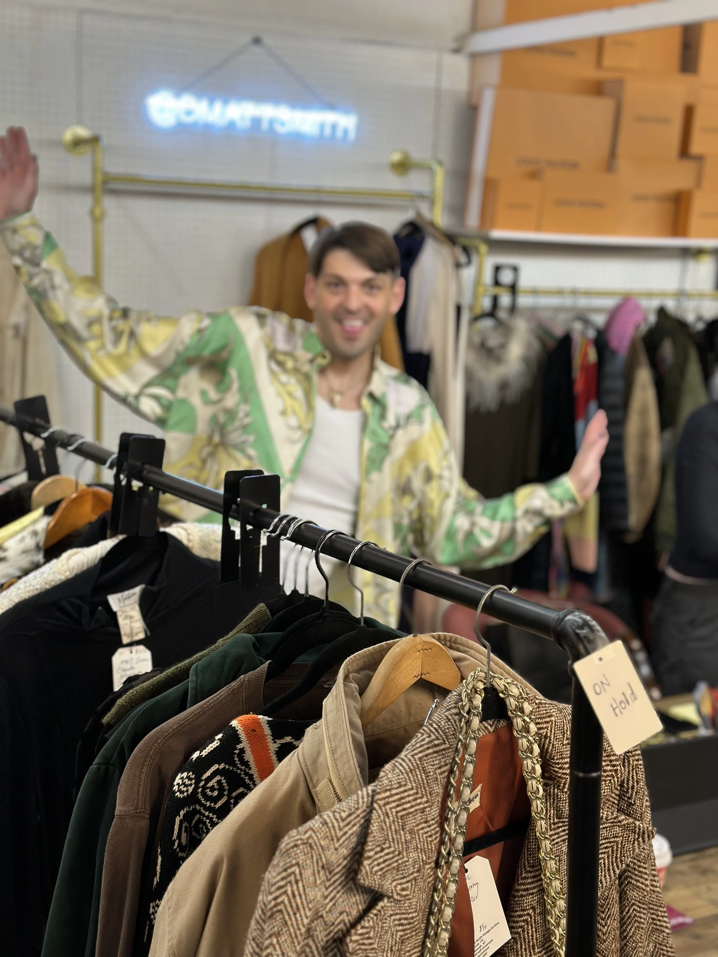 A person in a colorful patterned shirt, smiling and waving, standing behind a clothing rack with various jackets and shirts in a thrift store or clothing store. In the background, there are shelves with boxes and a blurred neon sign that reads 'GOMAT