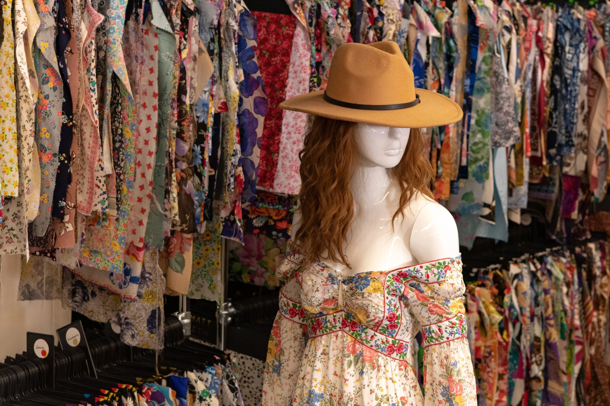 Mannequin with long, curly hair wearing a tan wide-brimmed hat and a colorful floral dress in a clothing store with racks of floral dresses in the background.