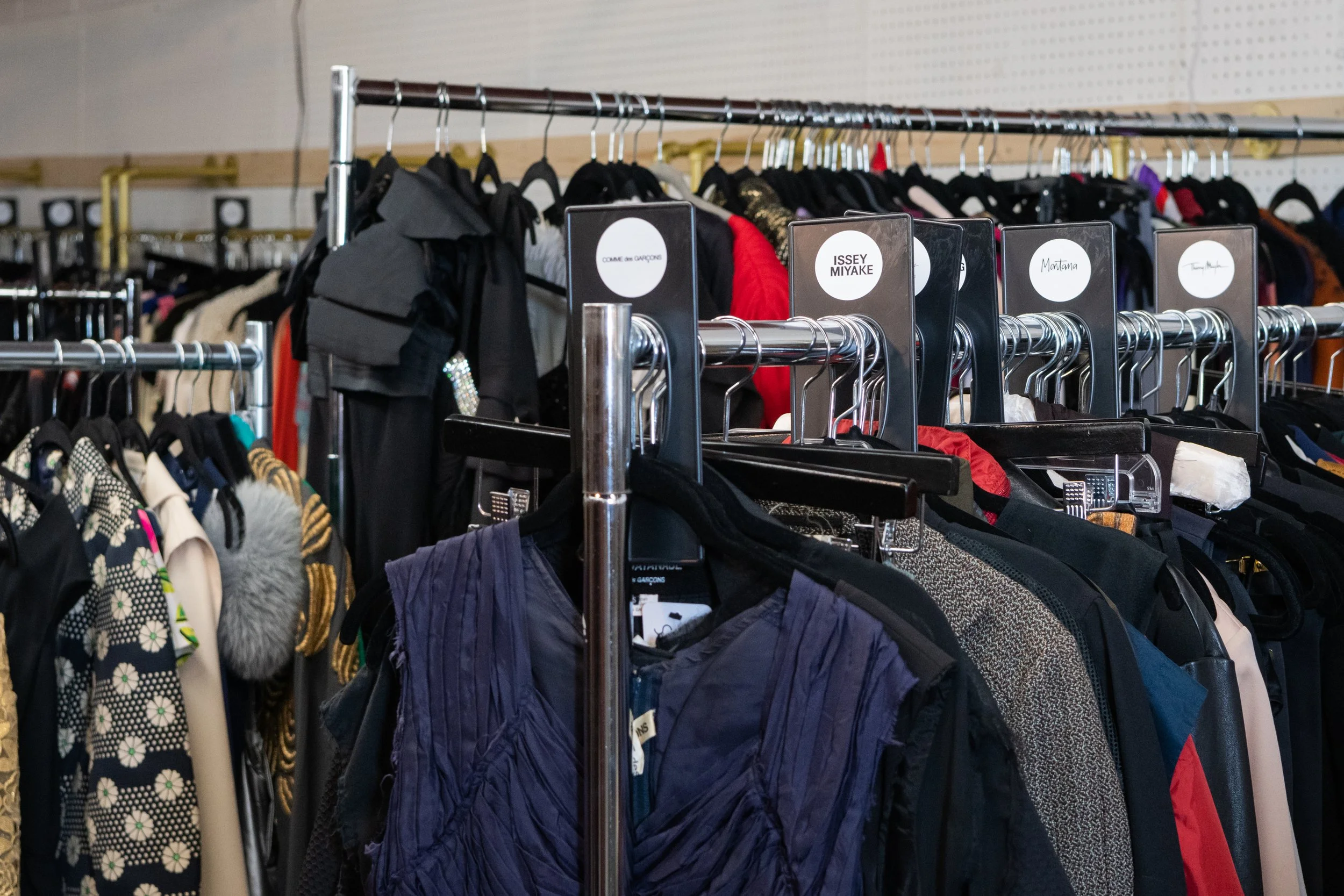 Clothing store with racks of various jackets, coats, and garments on black hangers, labeled with brand tags like Comme des Garçons, Issey Miyake, Montana, and Tom Ford.