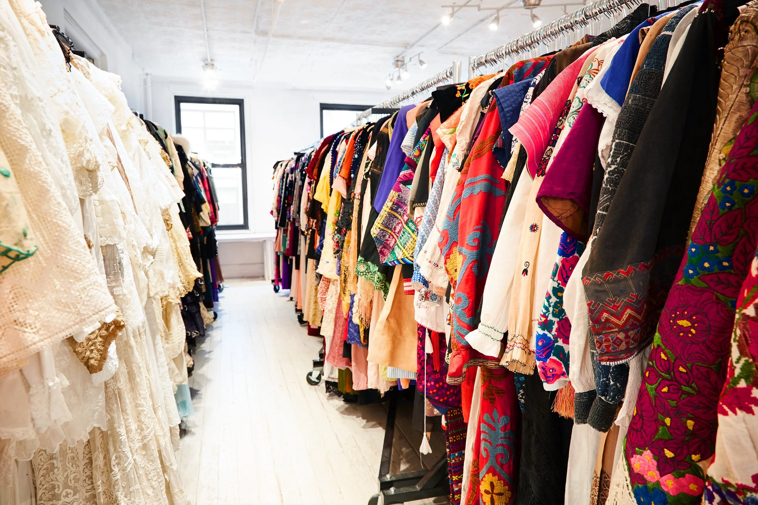 Clothing store with racks of colorful embroidered and textured garments, white walls, large windows, and overhead lighting.