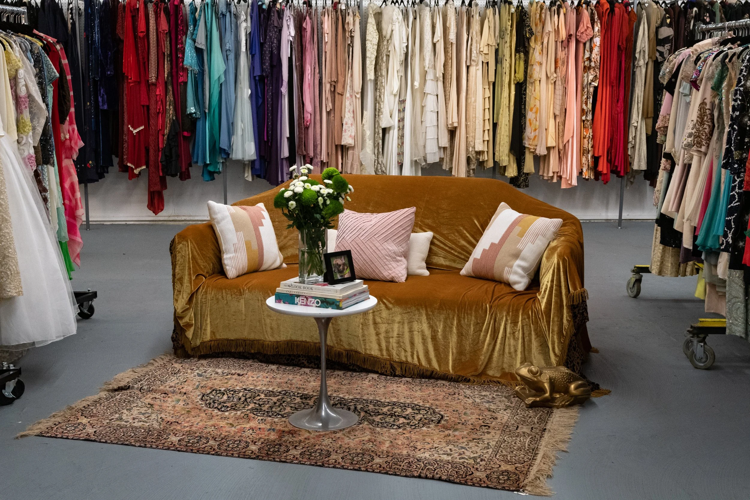 Inside a dress shop with clothing racks filled with colorful dresses, a velvet sofa decorated with pillows, a small table with flowers and books, and a patterned rug on the floor.