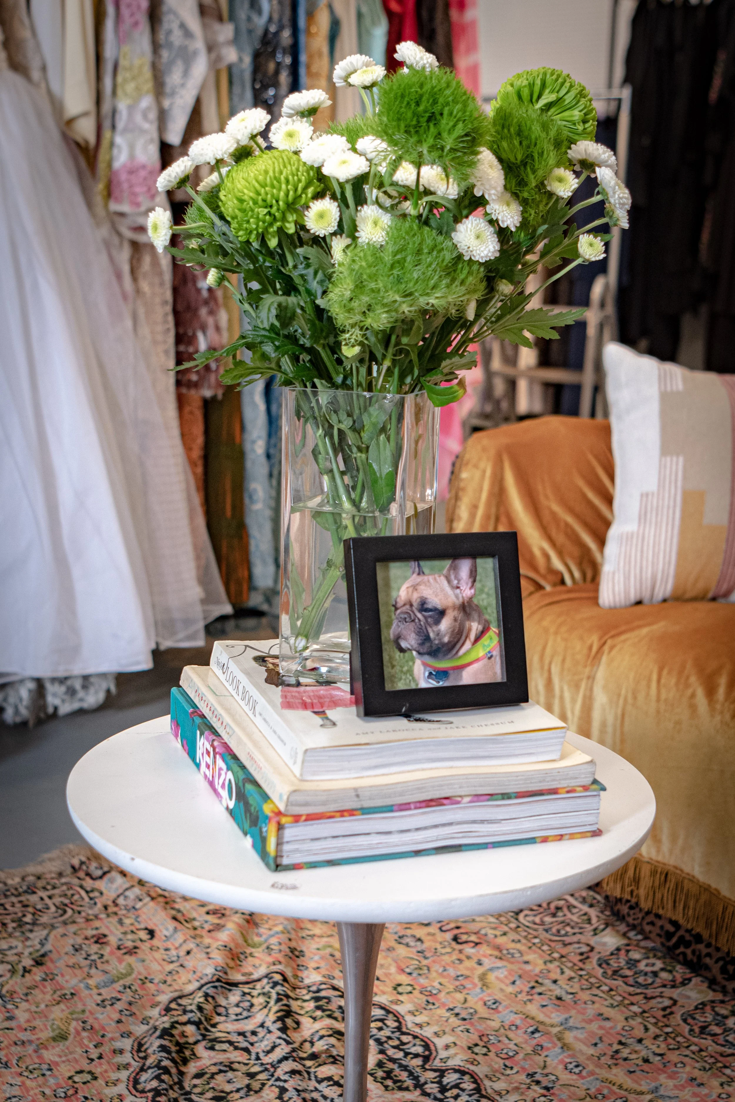 A vase of white and green flowers on a round white table, with a framed photo of a dog, and stacks of colorful books underneath. The background includes a patterned rug and a cozy living room setting with pillows and draped clothing.
