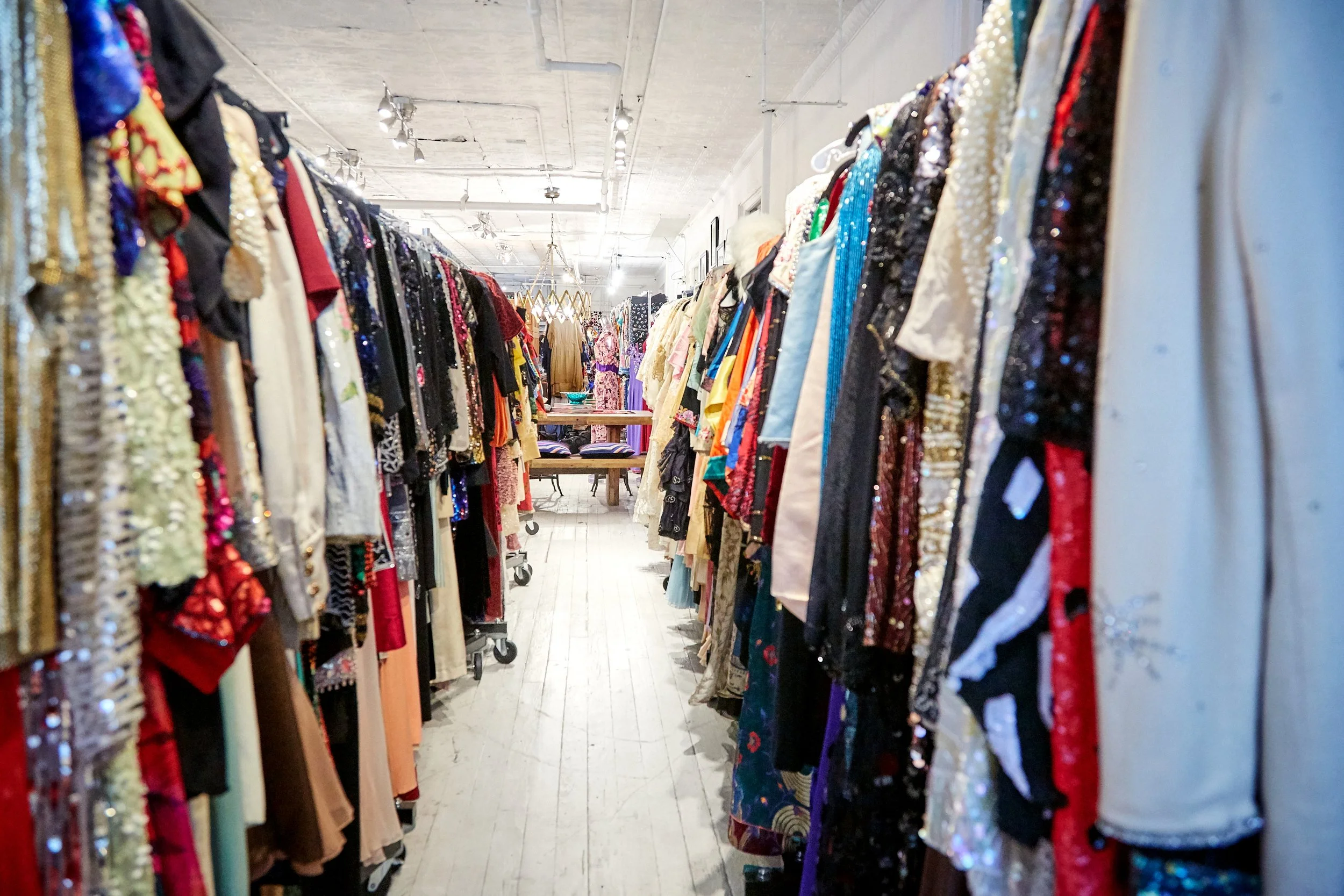 Midsection view of a vintage clothing store with racks of colorful dresses and garments on both sides, and a wooden table in the background, with a white ceiling and exposed industrial lighting.