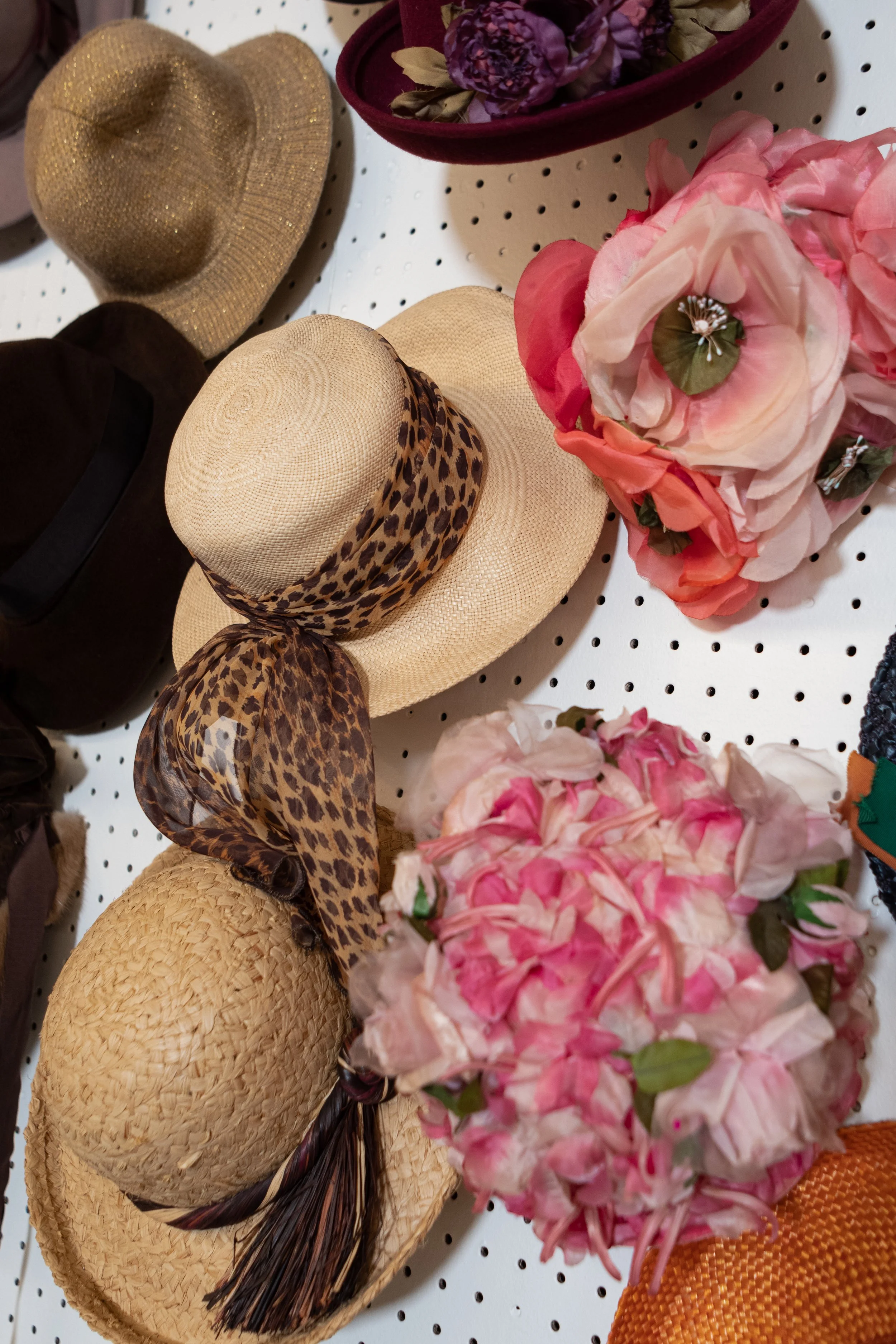 Assorted straw hats with different styles and patterns displayed on a white pegboard wall, including a beige woven hat with a leopard-print scarf, a black hat, a glittery gold hat, and a pink floral hat, along with pink and purple flower arrangements