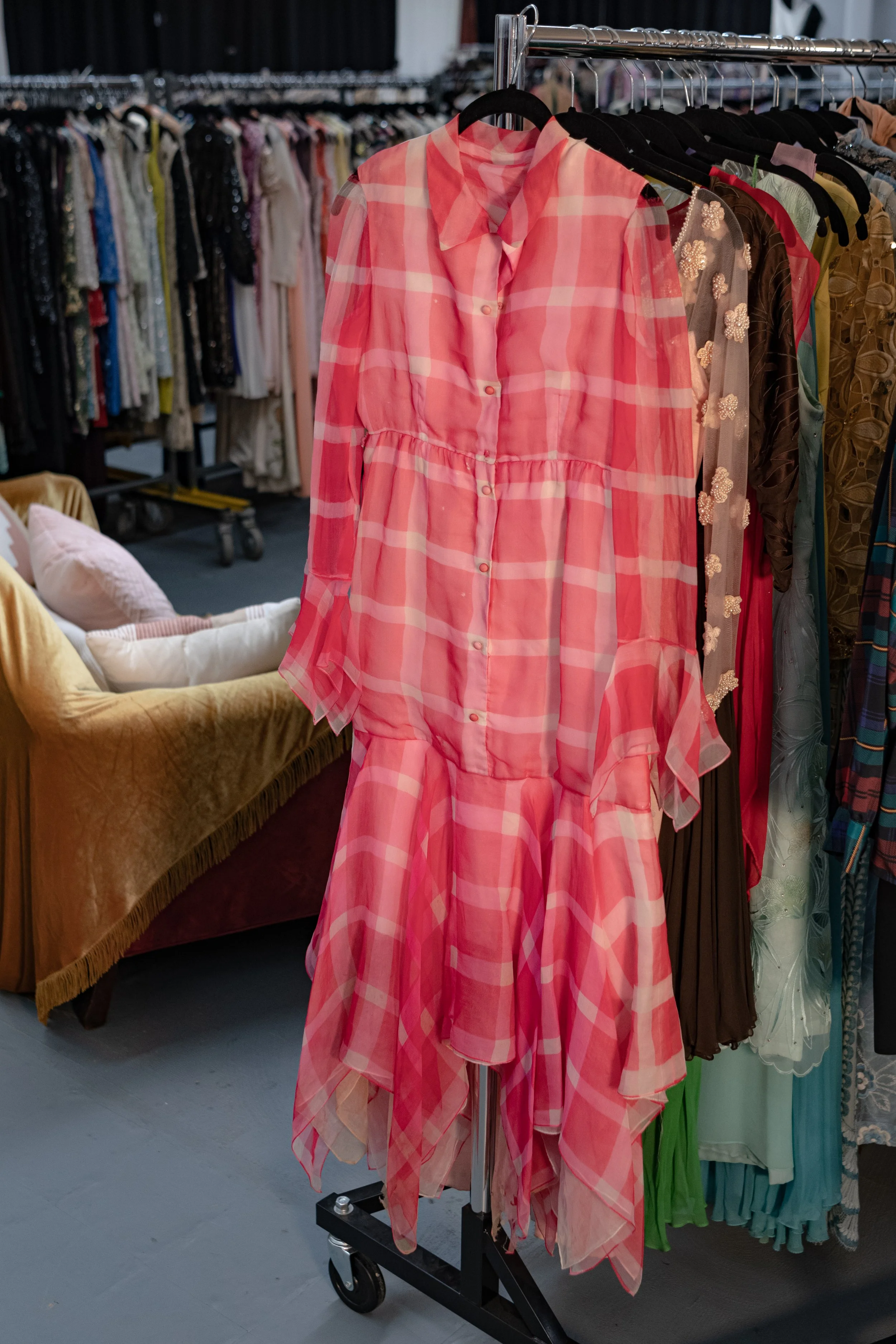 Pink and peach plaid dress hanging on a clothing rack in a thrift store or boutique. Other dresses are visible on the rack, and a yellow sofa with cushions can be seen in the background.