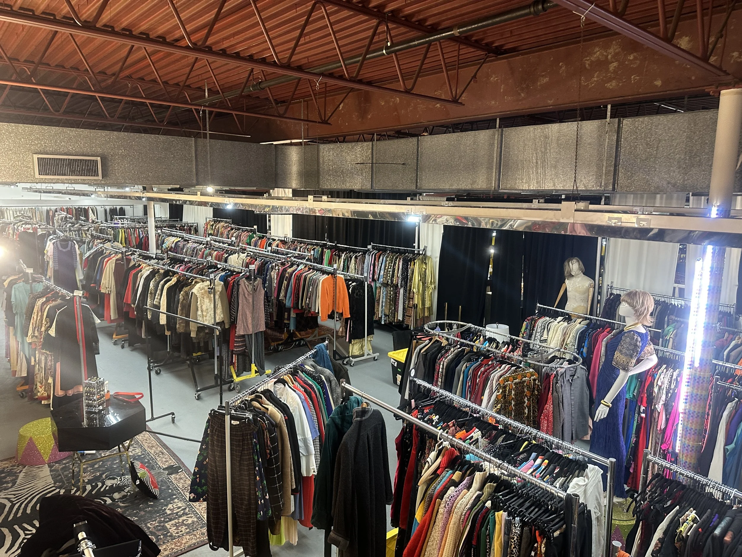 Clothing racks filled with a variety of colorful clothes inside a thrift or consignment store, with a patterned rug and mannequin dressed in a blue and gold garment.