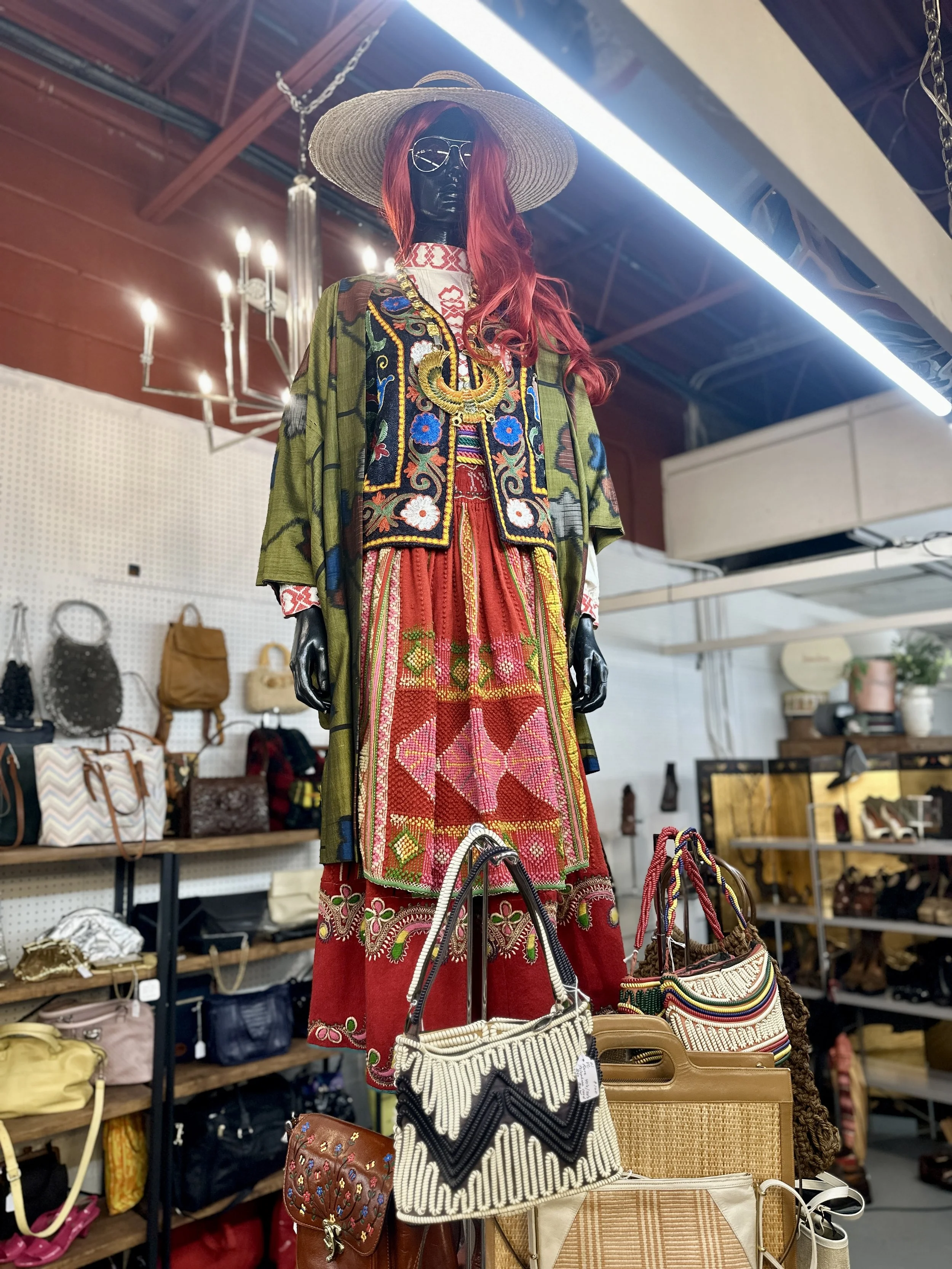 A mannequin dressed in a colorful, embroidered bohemian-style outfit, wearing a wide-brimmed hat and sunglasses, displayed in a boutique with shelves of handbags and accessories in the background.