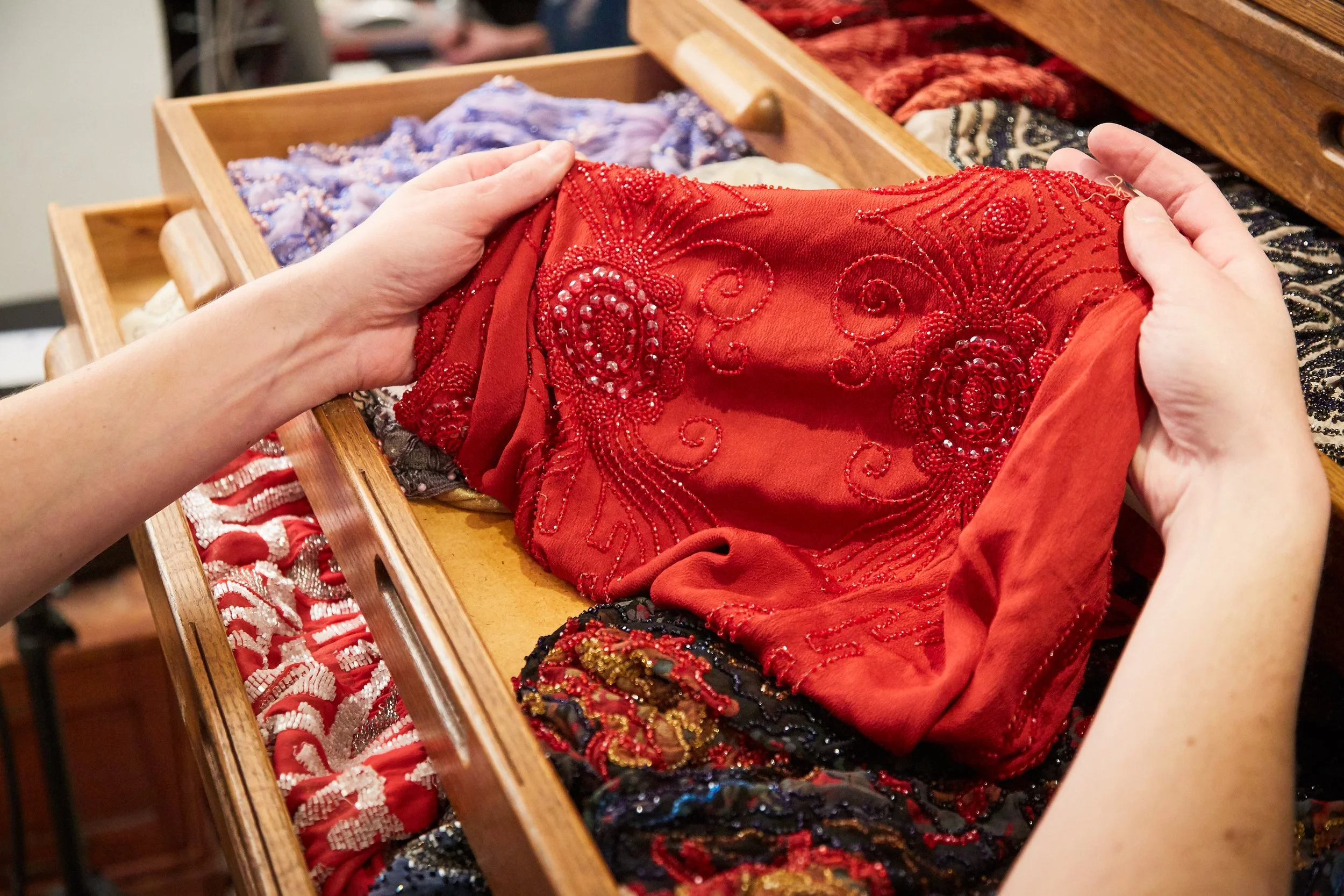 Hands holding a red, beaded, embroidered fabric with intricate floral patterns in a wooden box filled with various decorative fabrics and trims.
