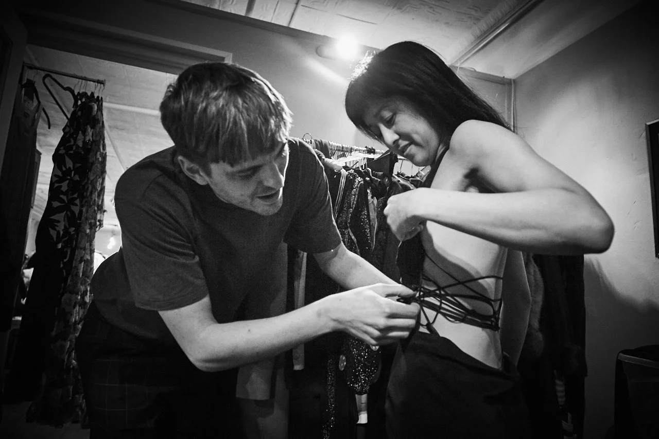 A woman is getting dressed with the help of a man, who is tying her outfit in a small room with clothing hanging in the background.