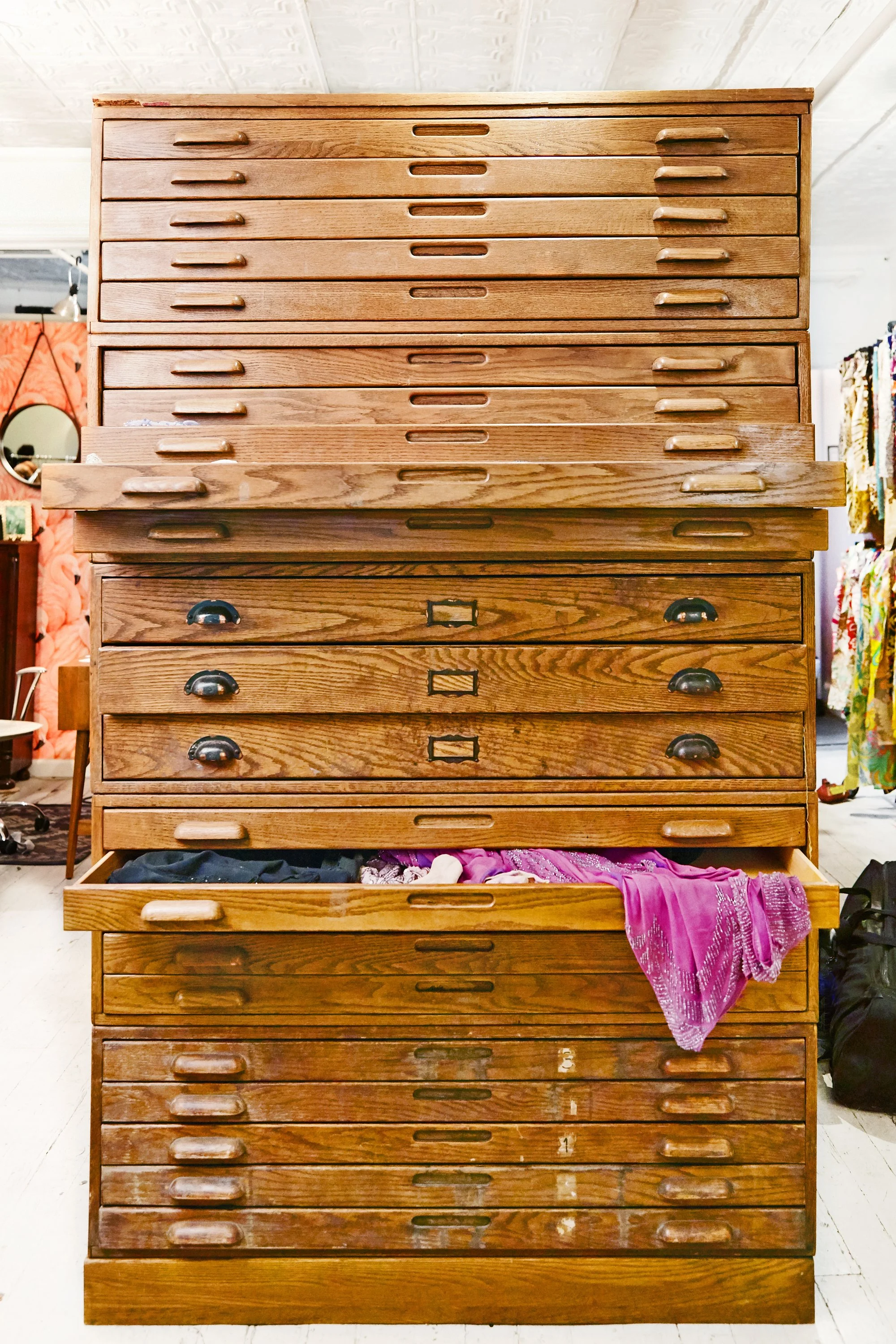 A tall wooden dresser with multiple drawers, some of which contain clothing and textiles, in a room with various decorations and furniture.