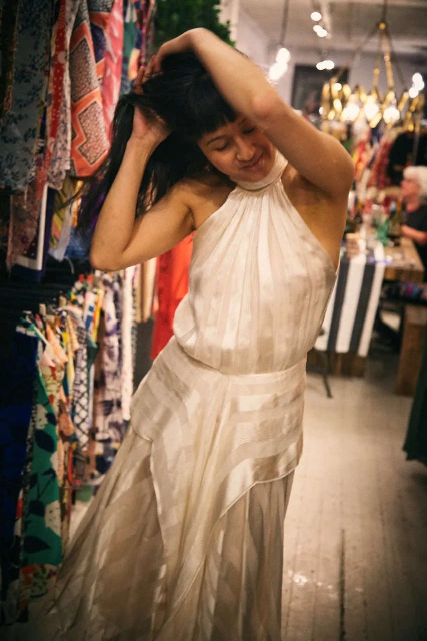 A woman with black hair wearing a shiny, cream-colored dress dancing in a boutique or market, with colorful fabrics and accessories displayed on racks and tables around her.