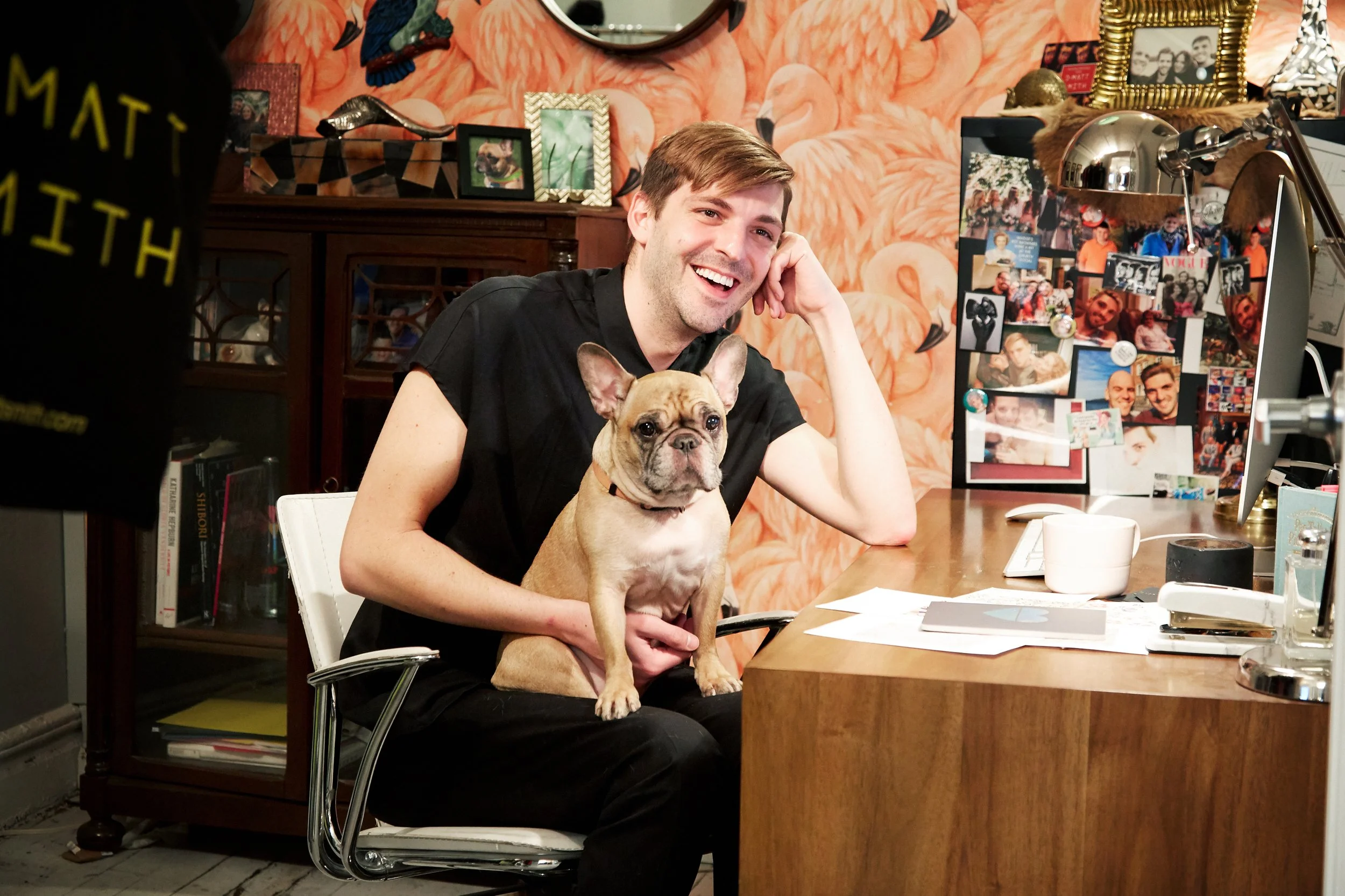 A young man sitting at a desk, smiling, holding a small dog, possibly a French Bulldog, on his lap. The background features a wall with a flamingo-themed wallpaper, decorative frames, and photos. The desk has papers, a lamp, and other personal items.