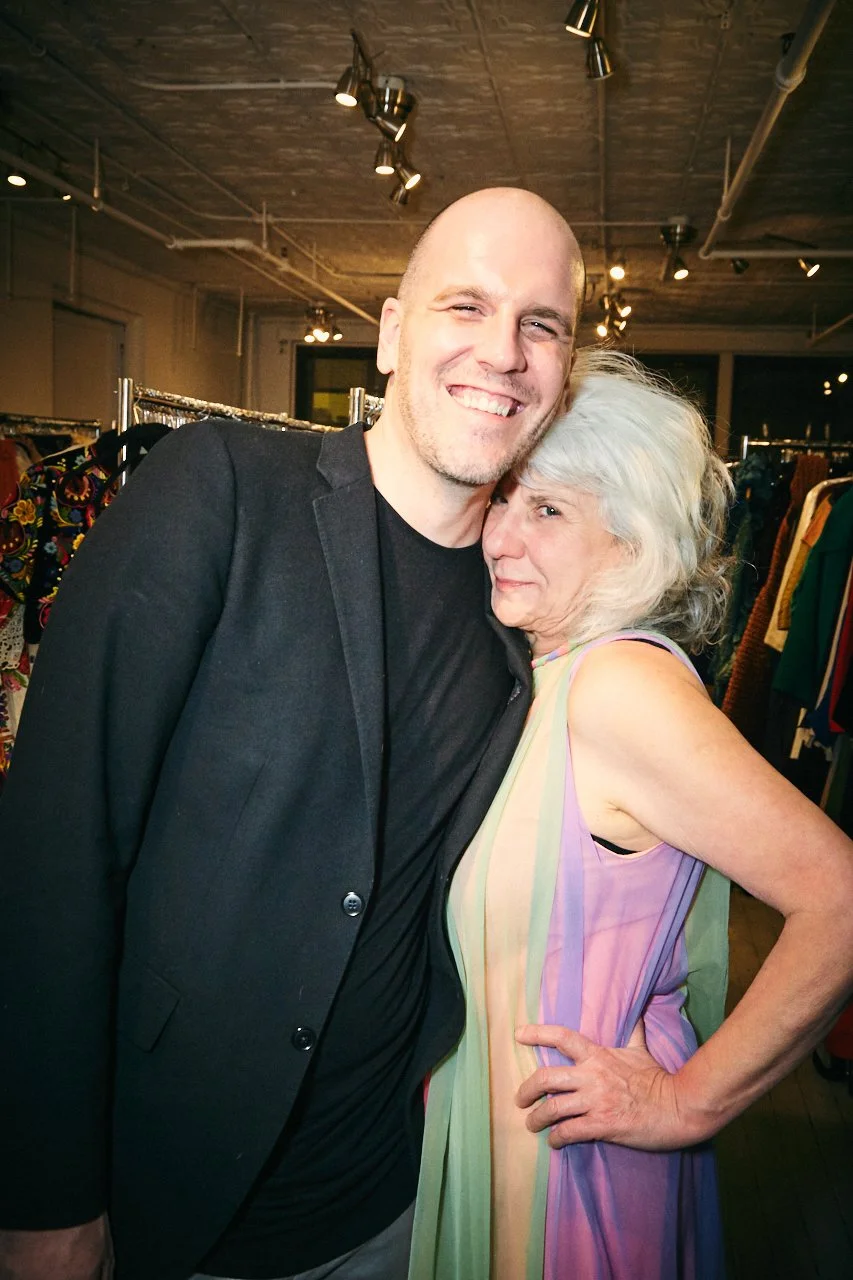 A man with a shaved head and a woman with white hair hugging and smiling at the camera inside a clothing store.