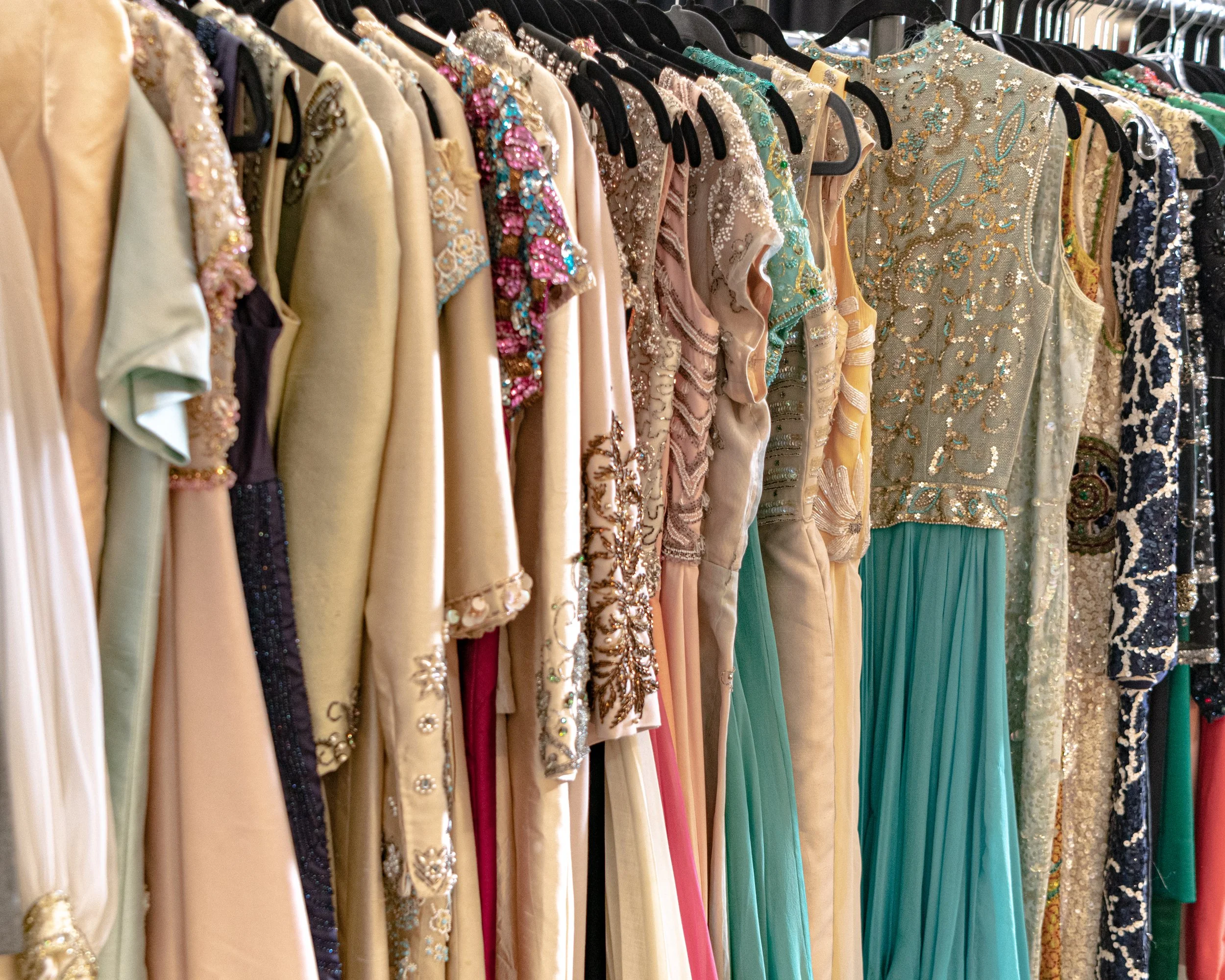 A row of colorful dresses hanging on black hangers, with elaborate embroidery and beading on some of the garments, in a retail clothing store.