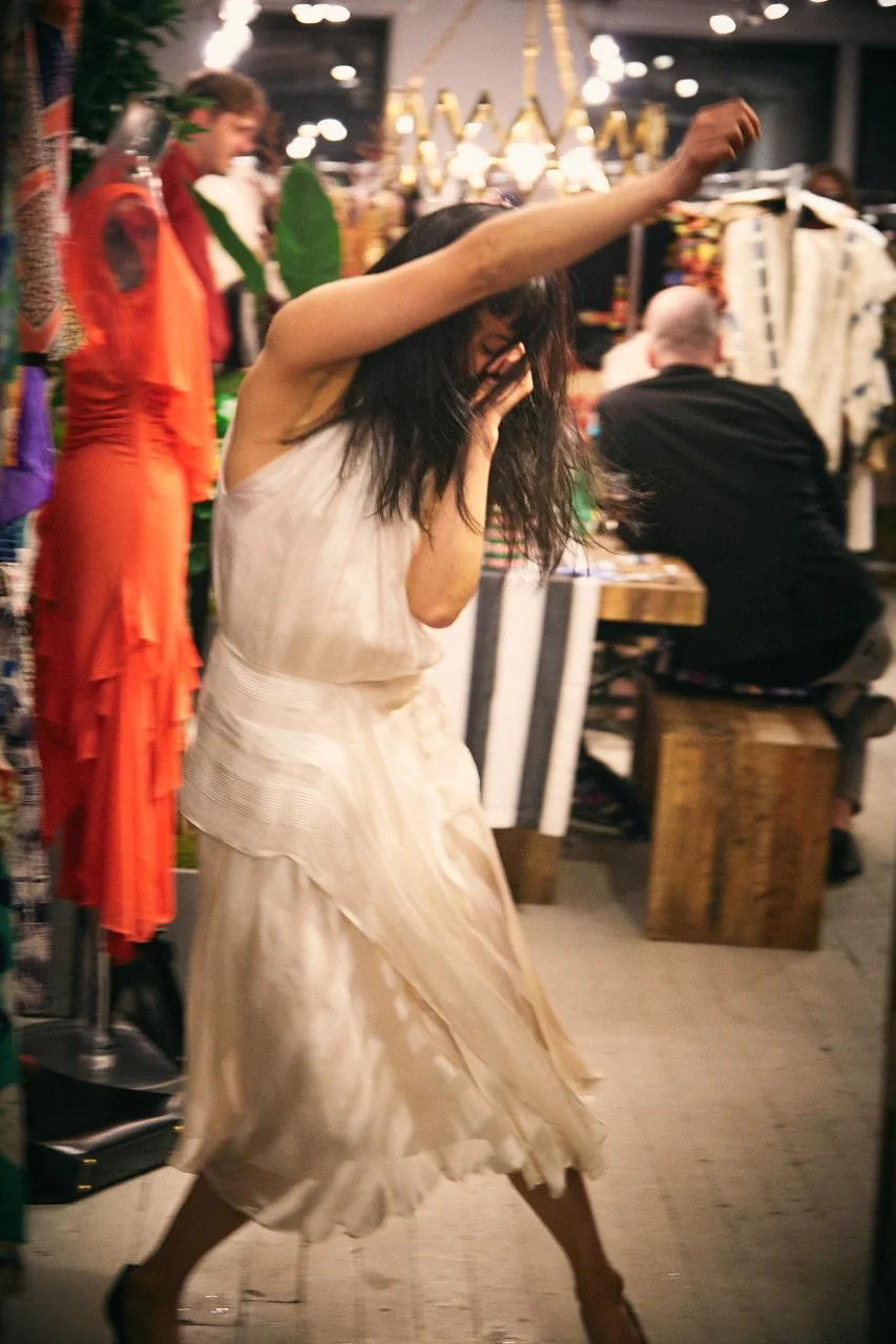 A woman in a white dress dancing with her hair covering her face in a lively store or market with colorful clothing and accessories around her.
