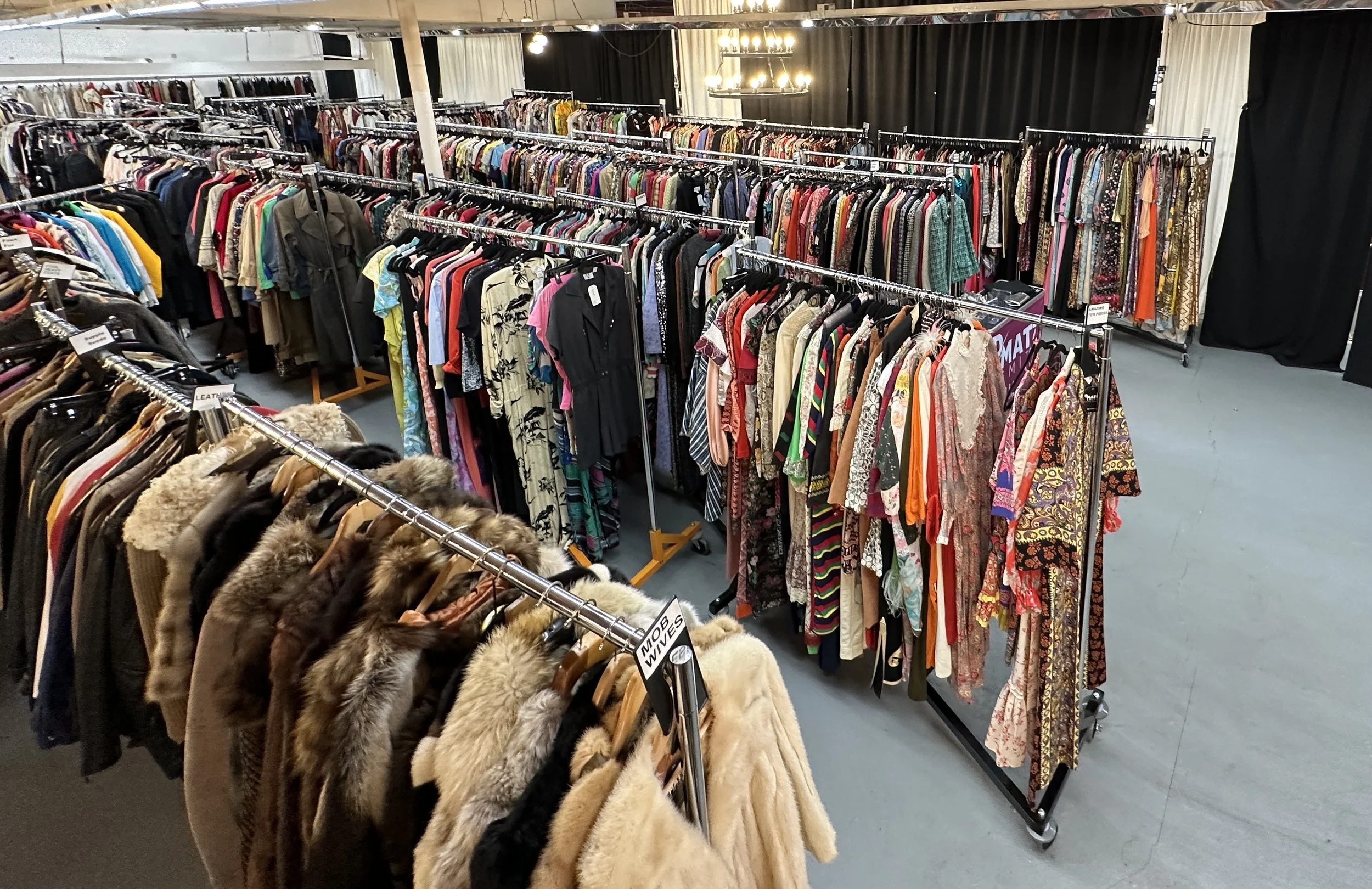 A clothing store with racks of dresses, shirts, and jackets. In the foreground, fur coats are hanging on a rack. The store has bright lighting and a black curtain backdrop.