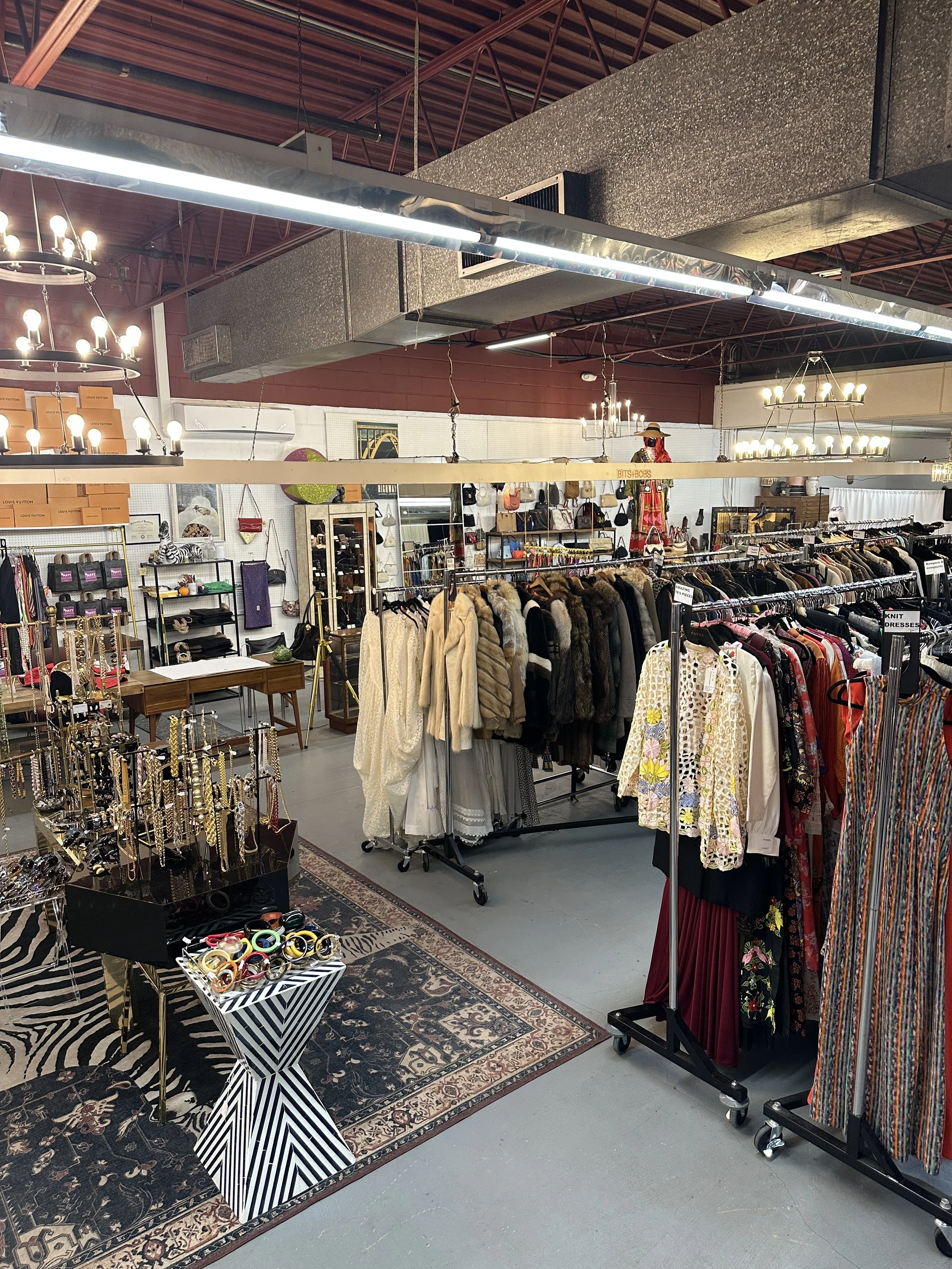 Interior of a vintage clothing store with racks of apparel, jewelry, handbags, and accessories, decorative lighting fixtures, and a patterned rug on the floor.