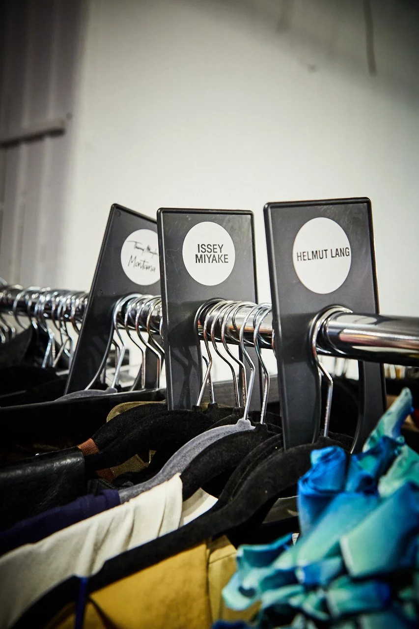 Clothing rack with hangers labeled with designer names, containing various garments like shirts and dresses.