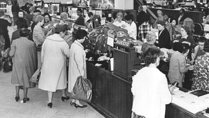People standing in line at a checkout counter in a store, with cashiers assisting customers.