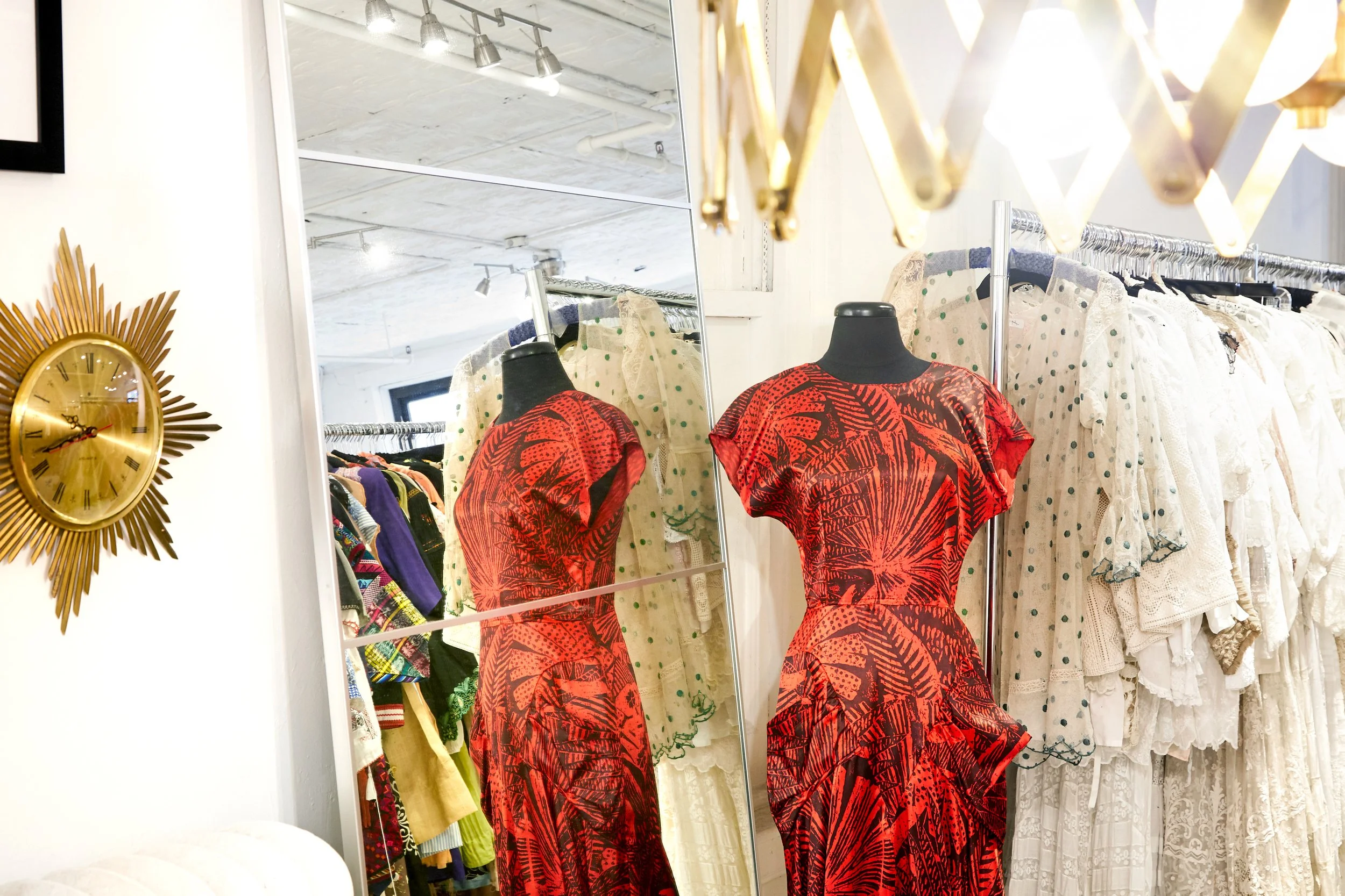 A clothing store displays two mannequins wearing red and black patterned dresses, with racks of various garments behind a mirror. A gold sunburst clock is on the white wall to the left.