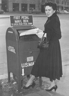 A woman in a dark dress and heels mailing a letter in a U.S. mail box labeled 'Foot Pedal Mail Box'.