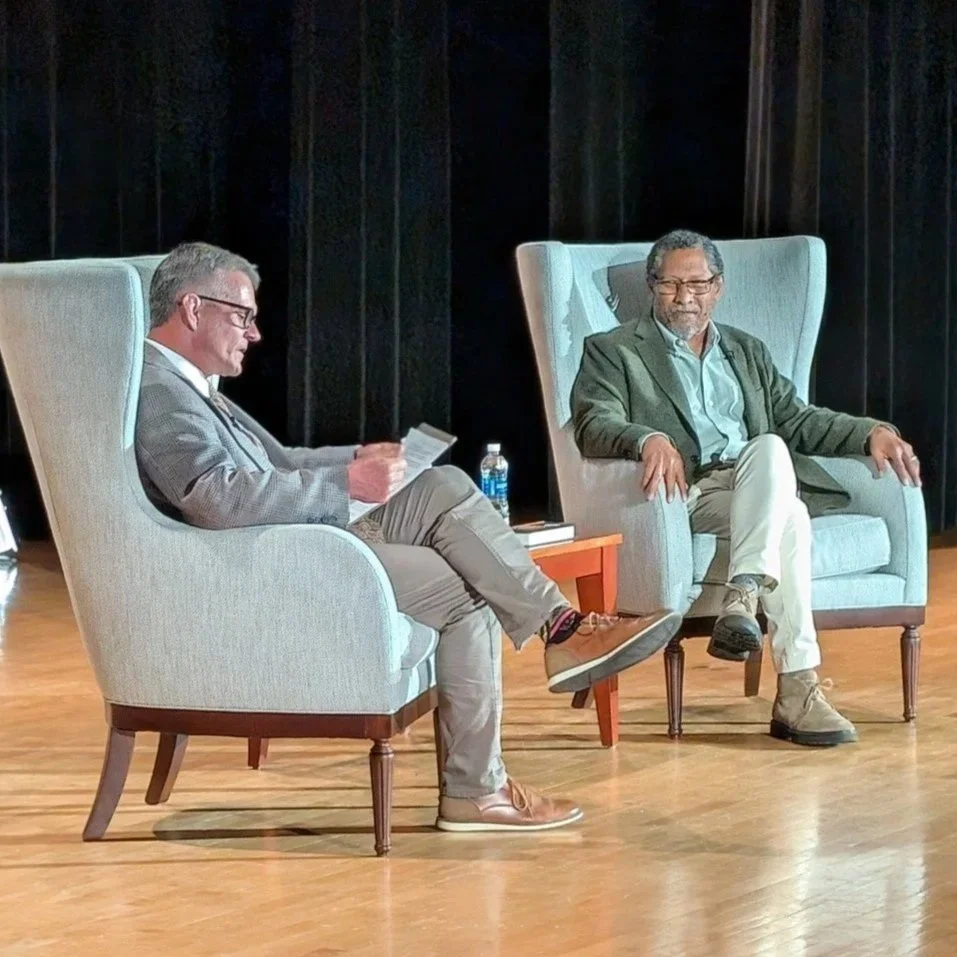 Two men sitting in armchairs on stage, engaged in a discussion. One man is reading from papers, and the other is listening attentively.