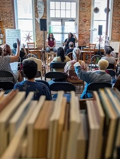 A panel discussion with three women on stage in a brick-walled room with large windows, surrounded by an audience of diverse people seated and listening.