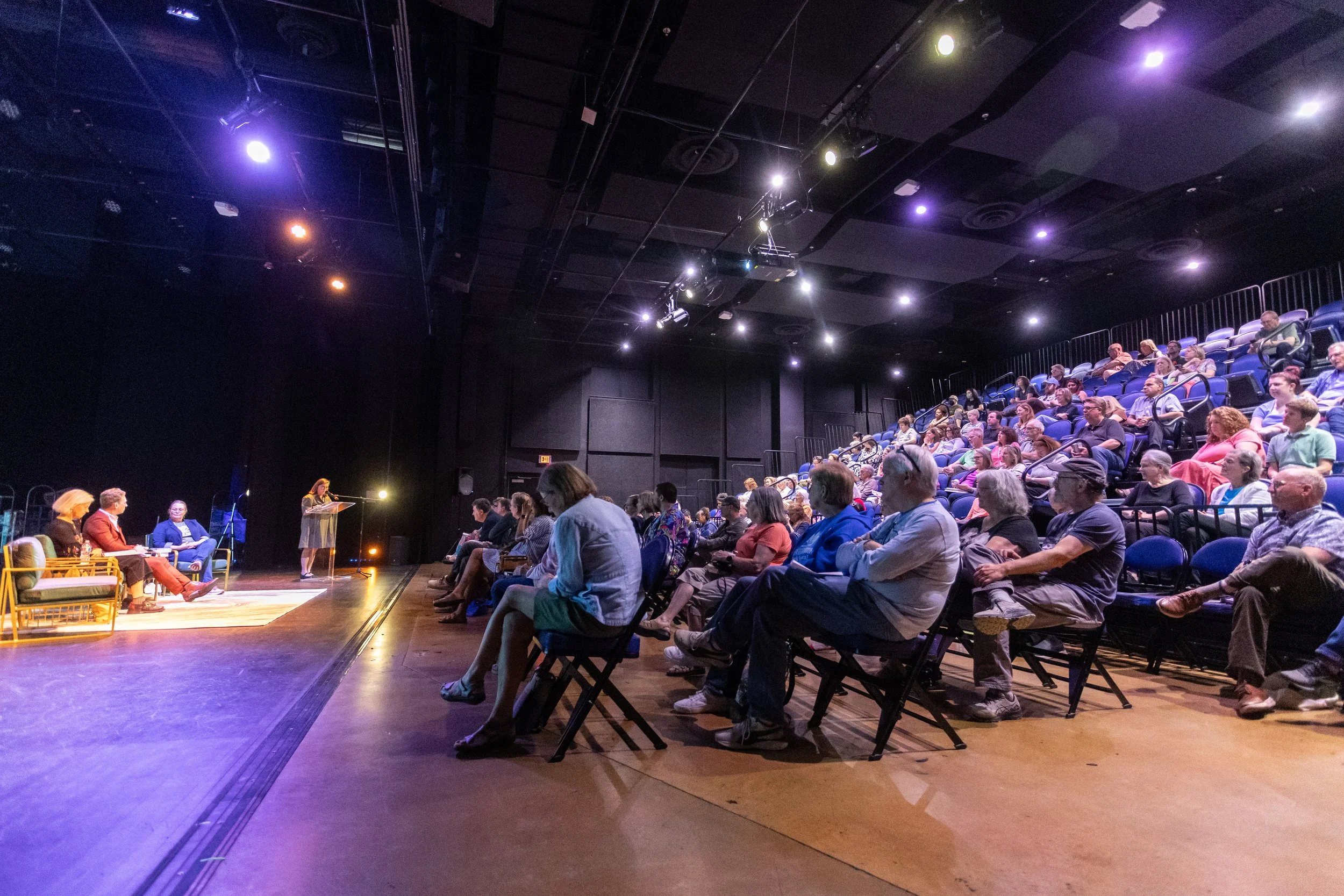 People attending a panel discussion or presentation in a theater or auditorium with a stage and seating area.