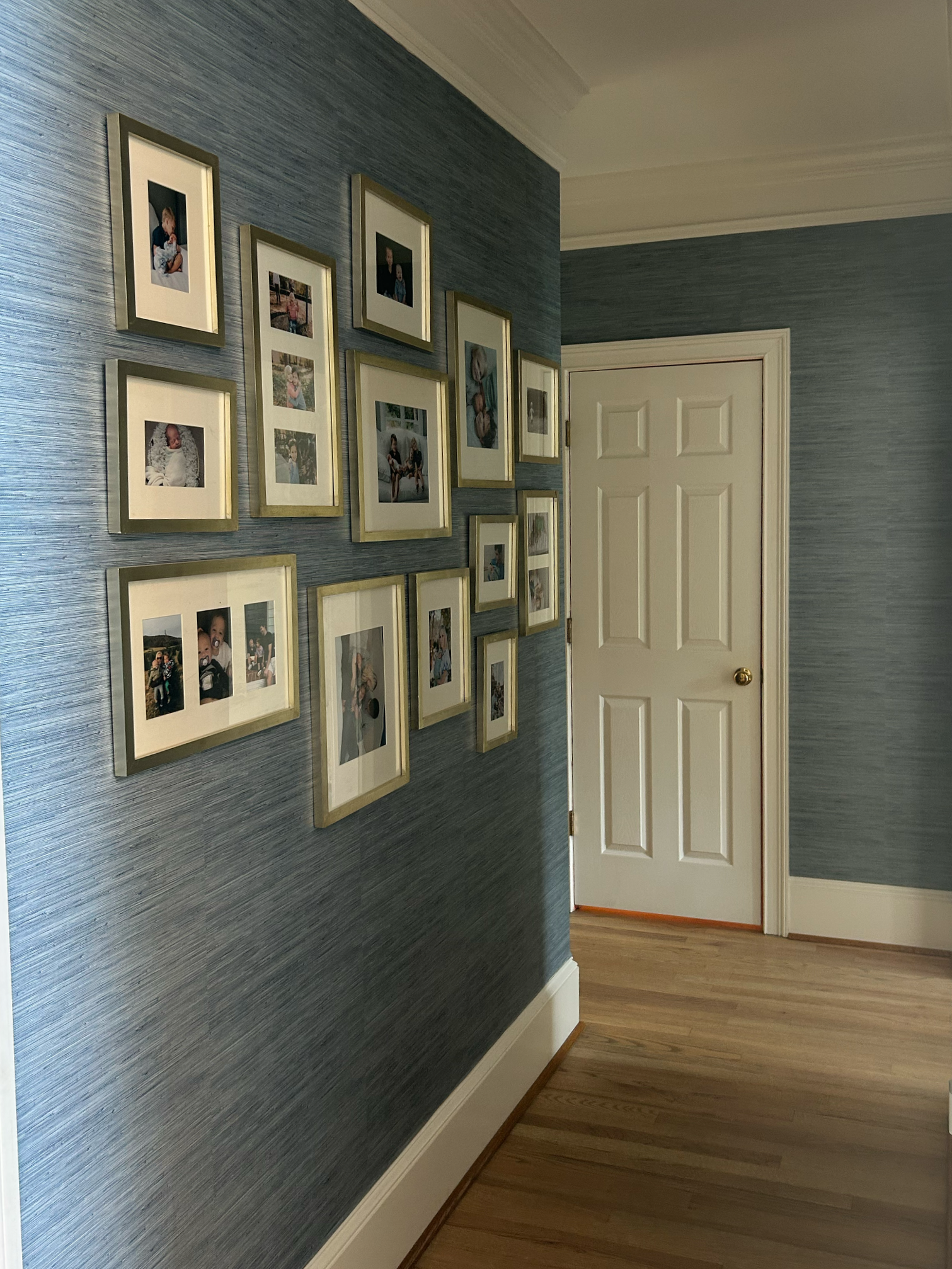 A wall with framed photographs arranged in a gallery style, adjacent to a white door with a gold-colored doorknob in a hallway with wood flooring and textured blue wallpaper.