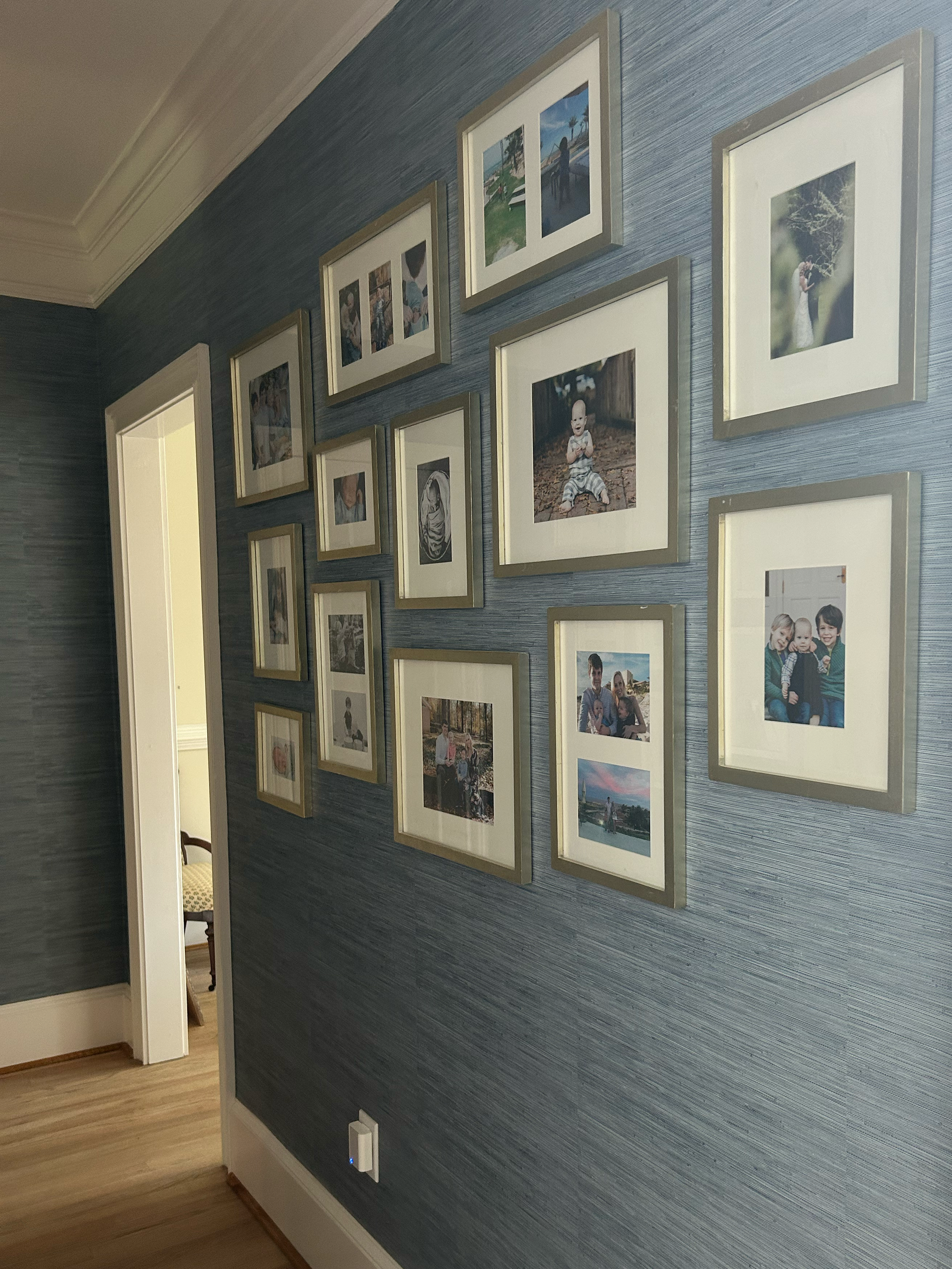 Photo of a blue textured wall decorated with a collection of framed family photographs arranged in a gallery style. Part of a doorway is visible on the left, leading to a room with a wooden chair.