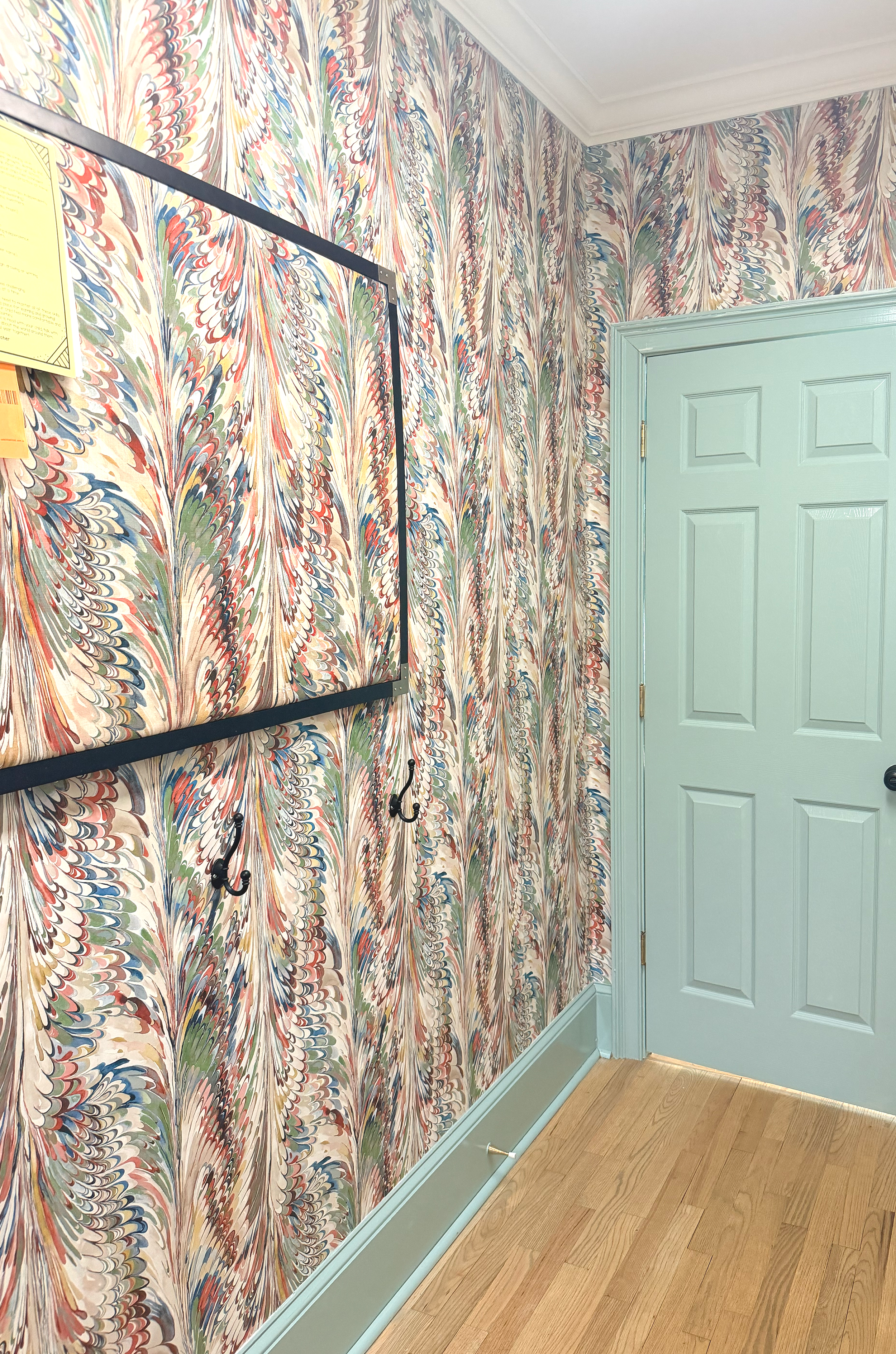 A wall decorated with colorful, marbled-pattern wallpaper next to a light blue door with molding, black hooks, and a wooden floor.