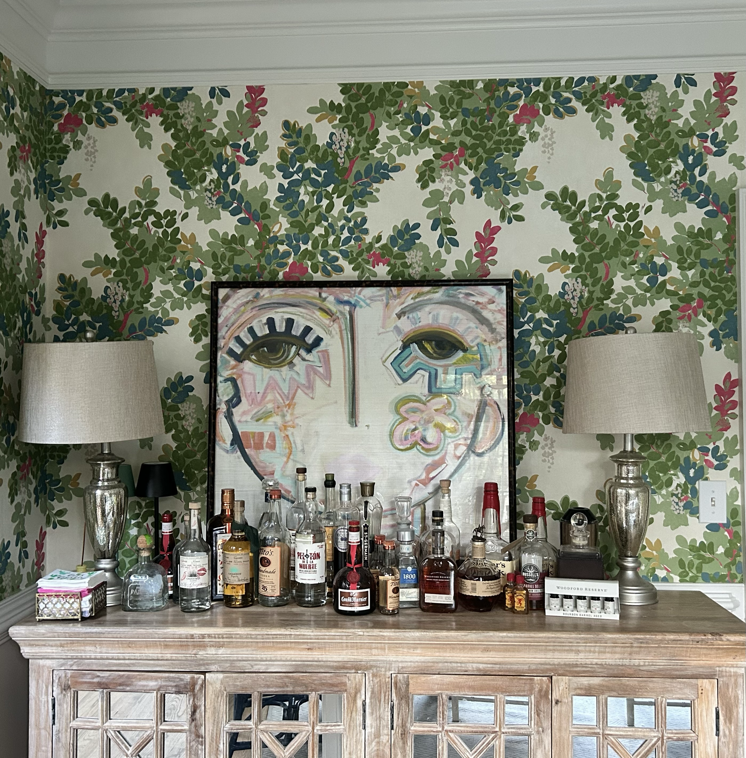 A wooden bar with assorted liquor bottles, two lamps, and a colorful artwork featuring eyes and abstract shapes on the wall behind it, with a leafy wallpaper pattern.