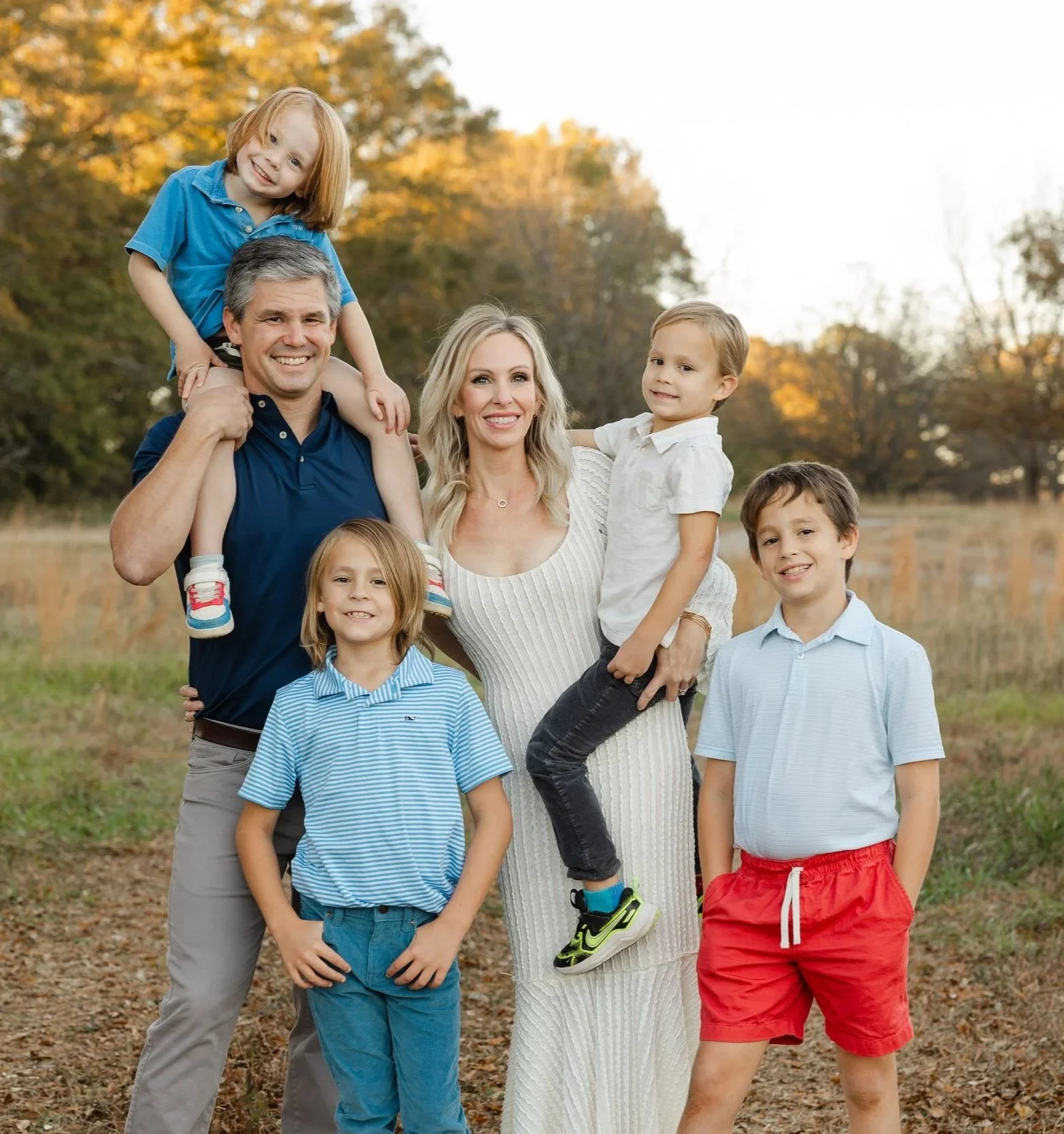A family of seven outdoors in fall landscape, two adults and five children, some children on shoulders, all smiling.