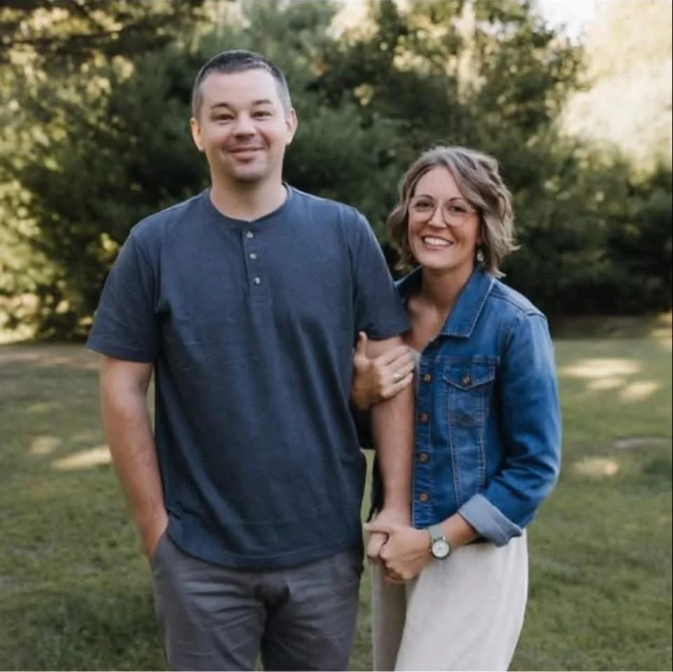 A man and woman standing outdoors in a park with trees in the background, smiling at the camera. The man has short dark hair, is wearing a blue T-shirt and gray pants. The woman has short, wavy hair, is wearing glasses, a denim jacket, and light-colored pants.