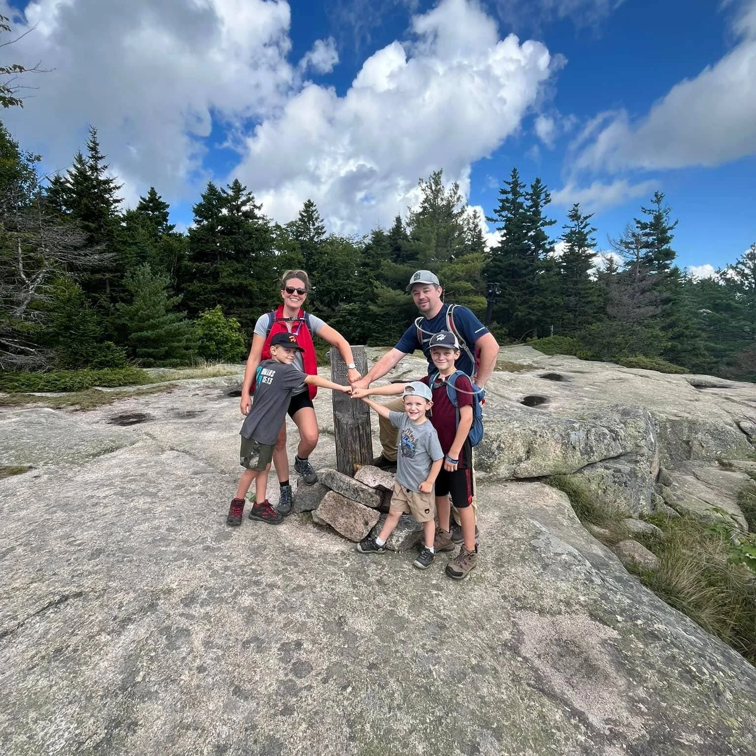 A family of five, including a woman and four children, standing on a rocky trail in a forest, holding hands around a wooden marker post with rocks at its base, under a partly cloudy sky.