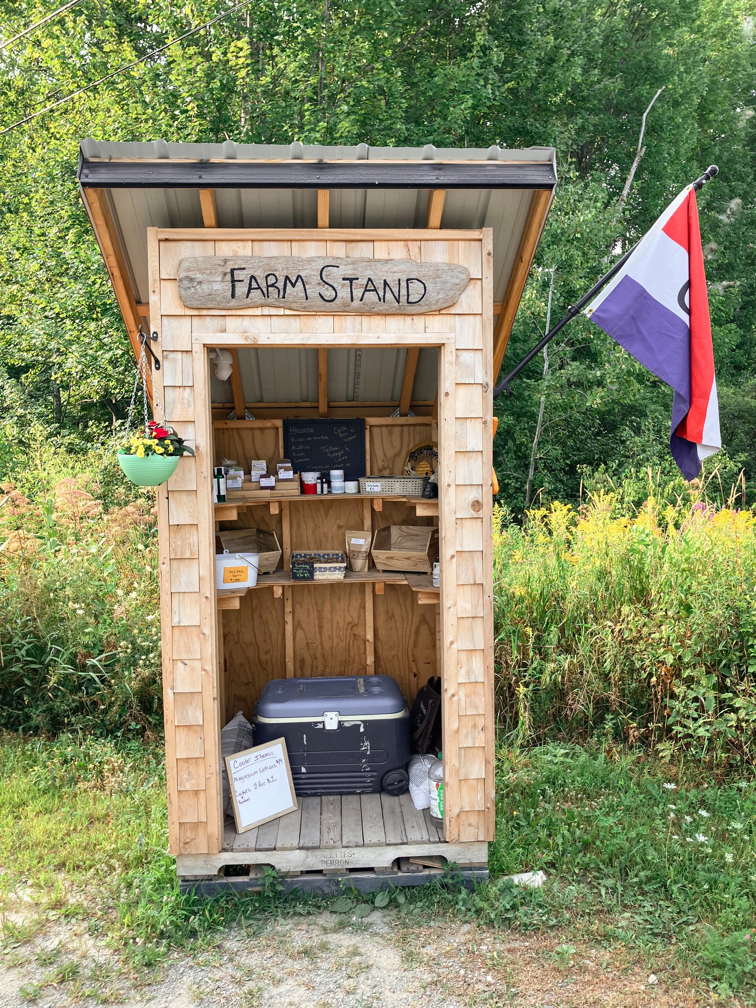 A small wooden farm stand with a sign that says 'Farm Stand', located outdoors among green trees and bushes. The stand has shelves with various jars and containers, a hanging flower pot, a Colorado flag on a pole, and a cooler on the floor.