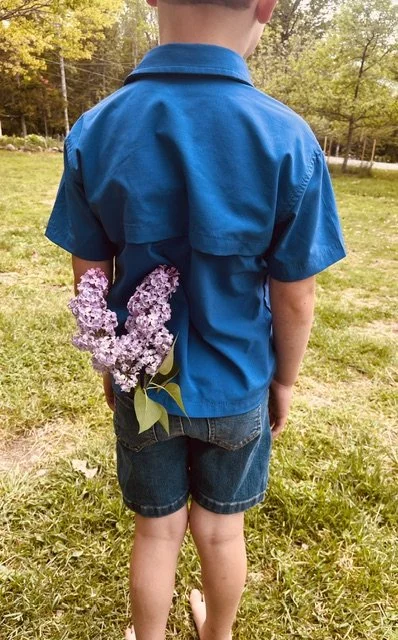 A child standing outdoors wearing a blue shirt and denim shorts, with a bunch of purple lilac flowers tucked into the back pocket of the shorts.