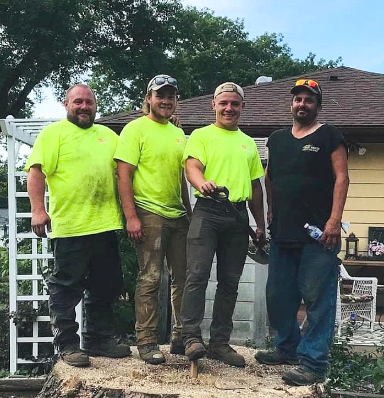 Four men standing outdoors on a tree stump, smiling at the camera, with a house and trees in the background.