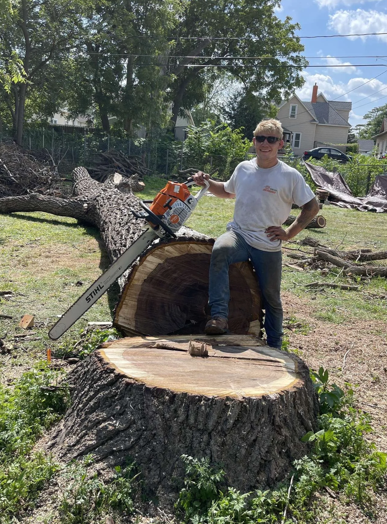 A young man wearing sunglasses, a white t-shirt, and ripped jeans stands on a large tree stump in a yard with cut trees and wood debris, holding a chainsaw.