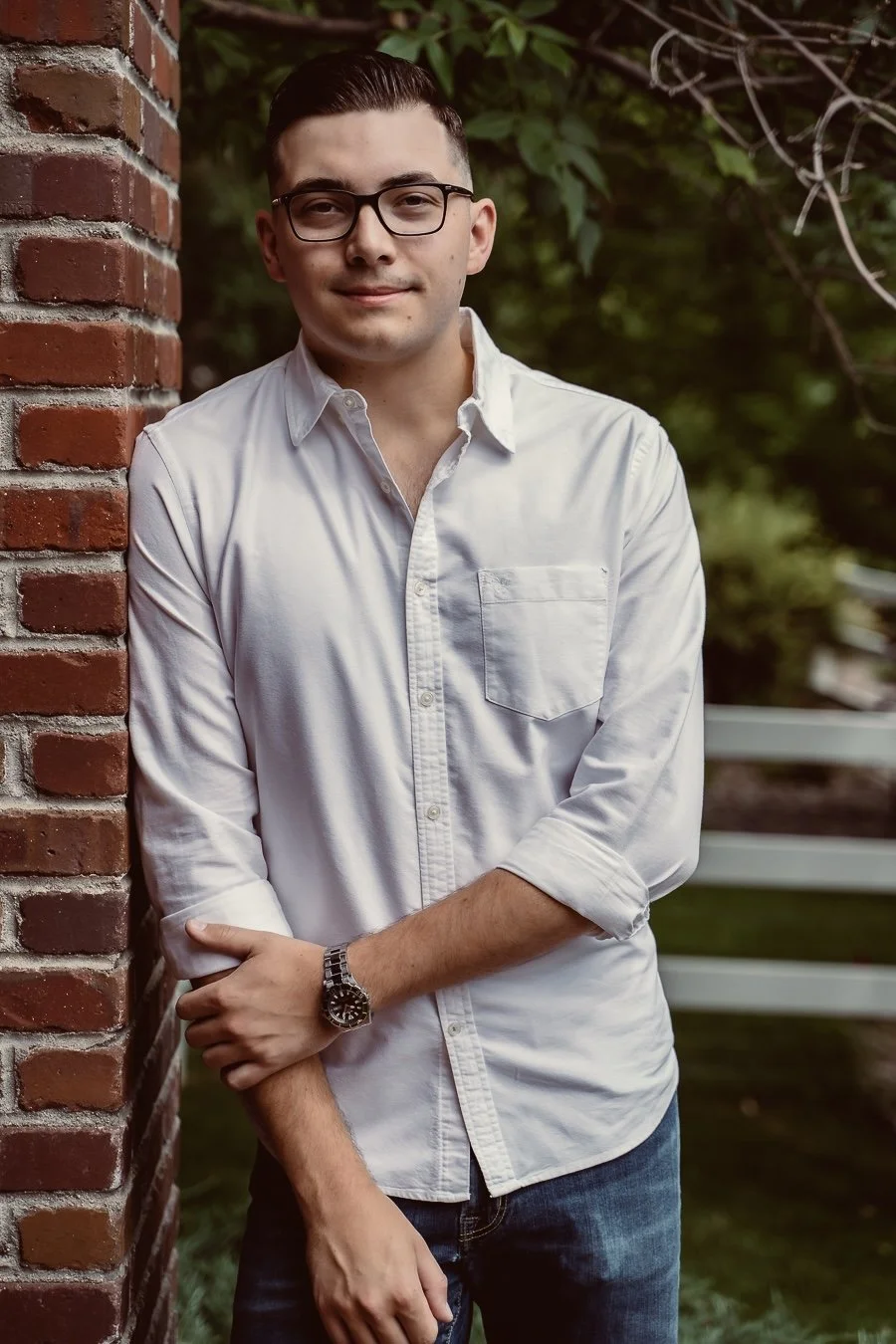 A young man with dark hair, glasses, wearing a white button-up shirt and jeans, standing outdoors next to a brick wall with greenery in the background.