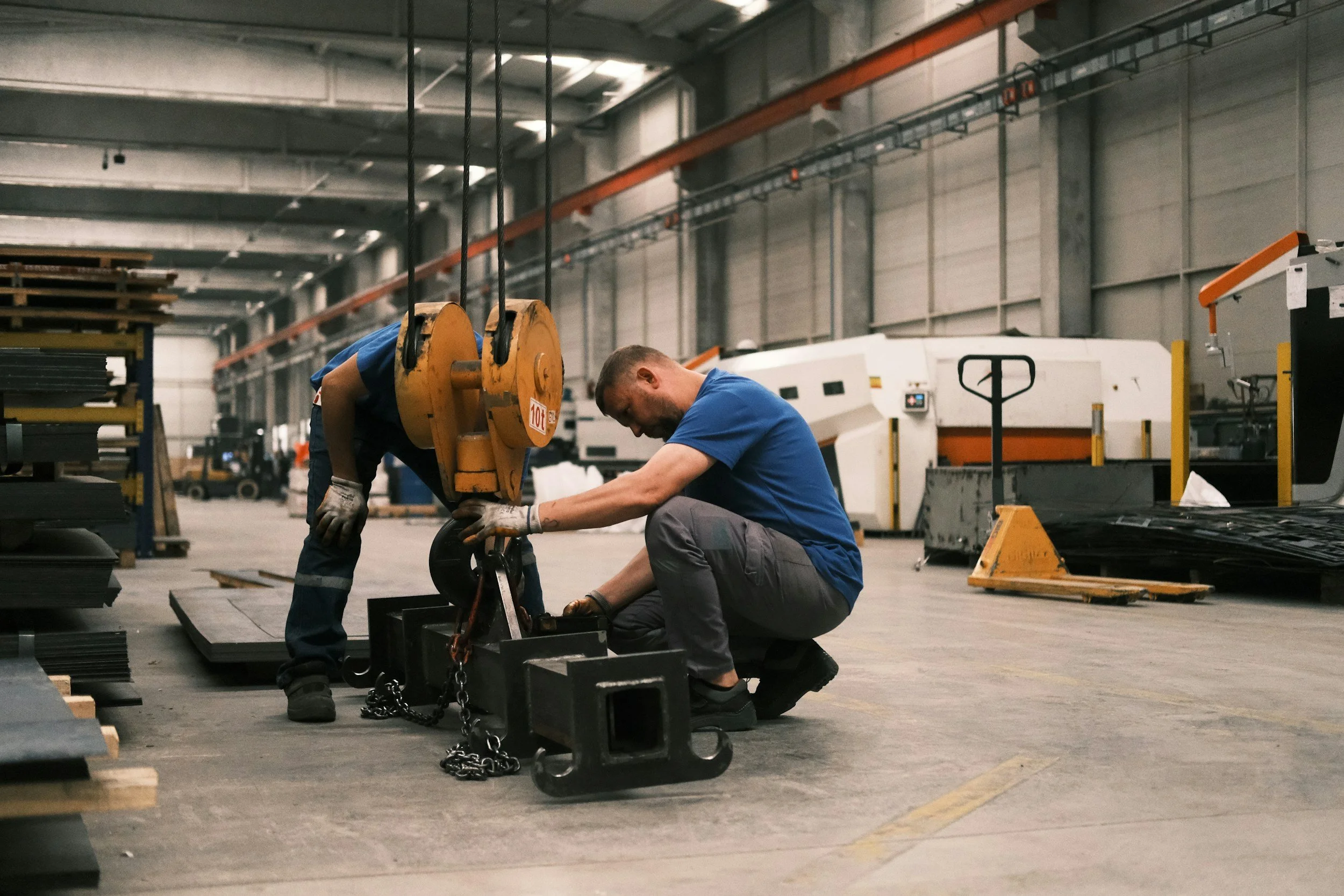Two workers in a factory or warehouse are assembling or repairing equipment. One worker is kneeling while the other is bent over, working together on a metal piece. The background shows industrial machinery and supplies, with a high ceiling and overhead lights.